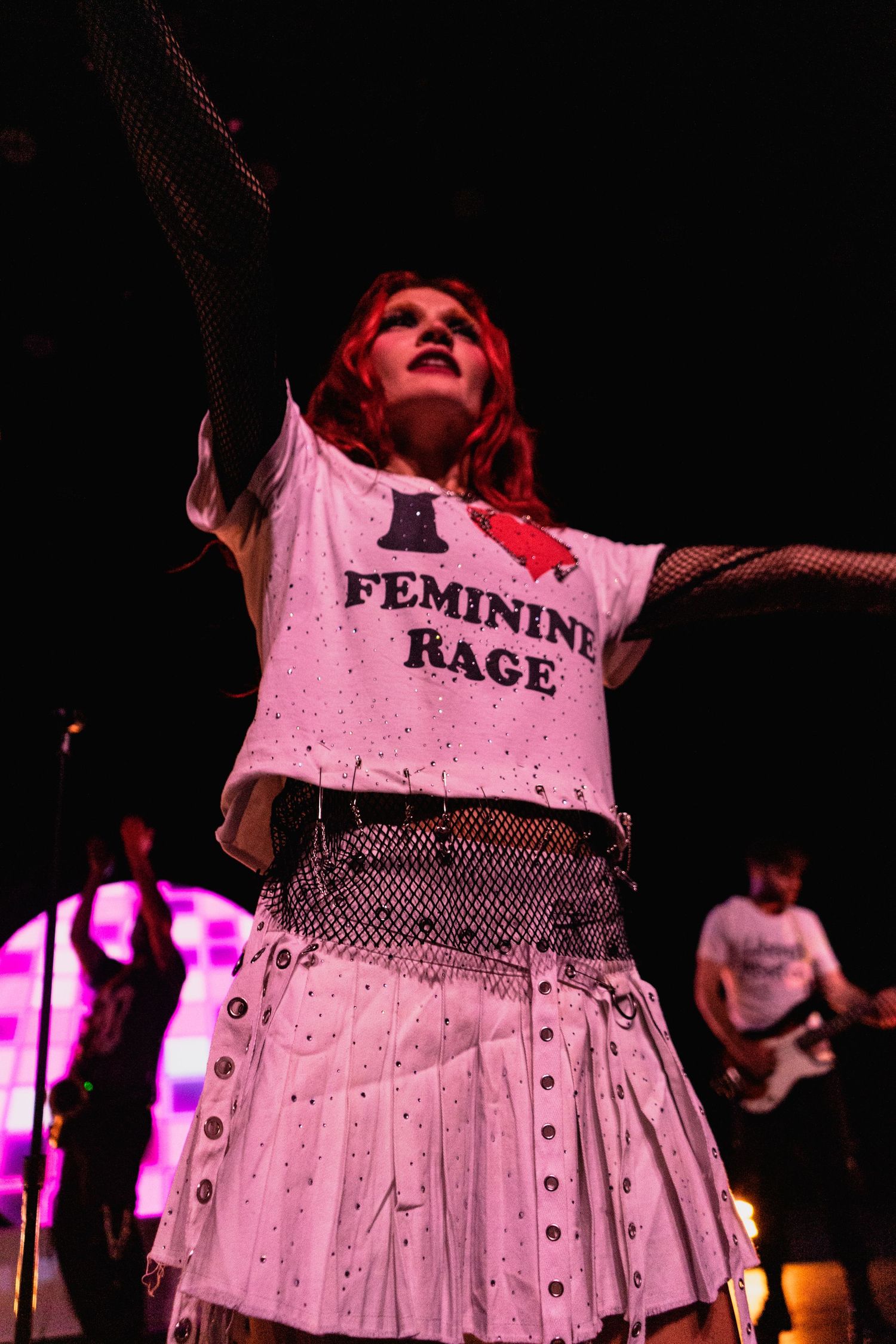 Female performer on stage wearing white Feminine Rage t-shirt with pleated skirt.