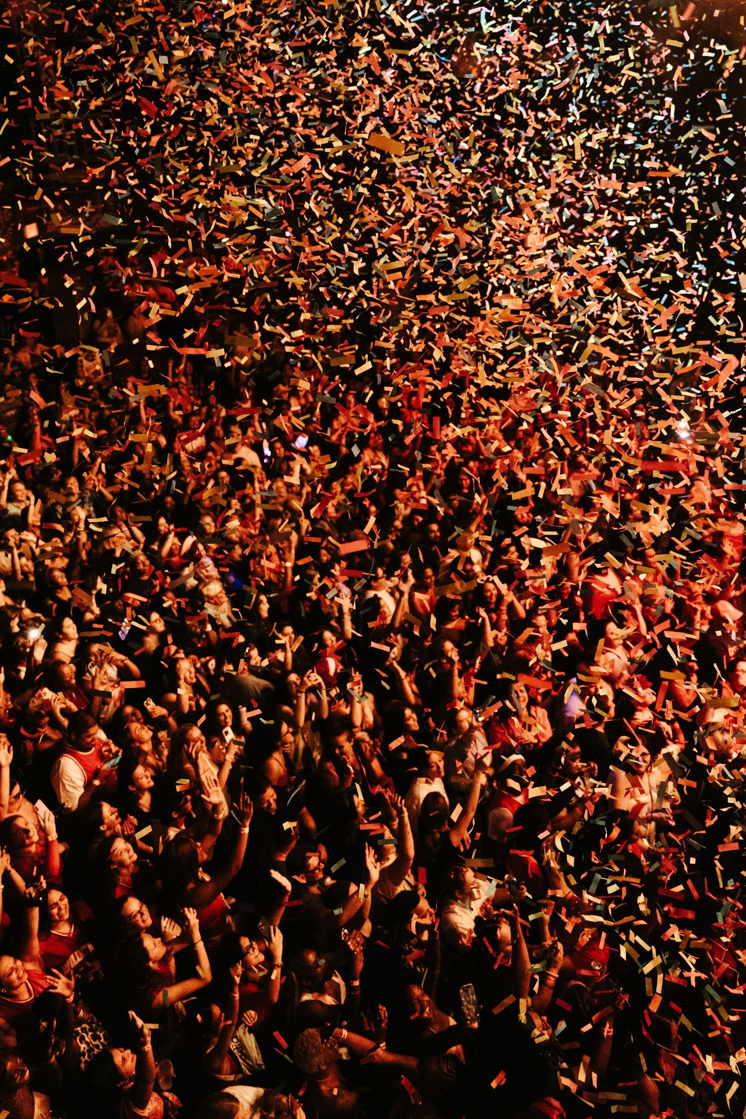 Massive concert crowd celebrating with confetti raining down under warm red and orange stage lighting.