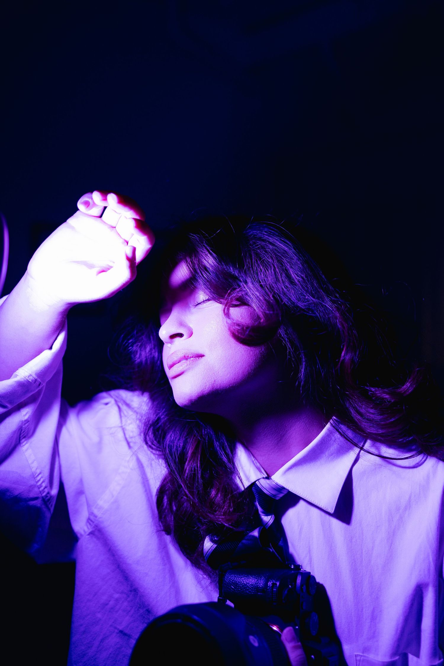 Artistic purple-lit portrait of a young person in white shirt and tie holding a camera in darkness.