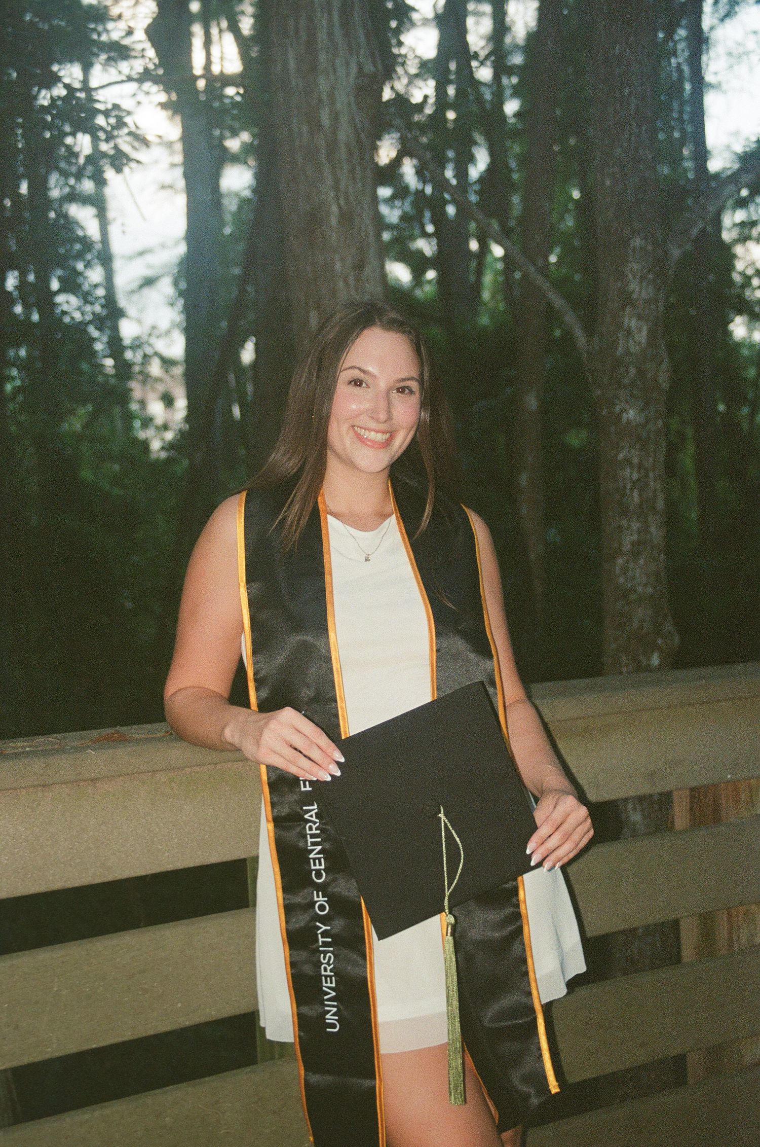 Smiling female UCF graduate holds cap while wearing stole outdoors among trees.