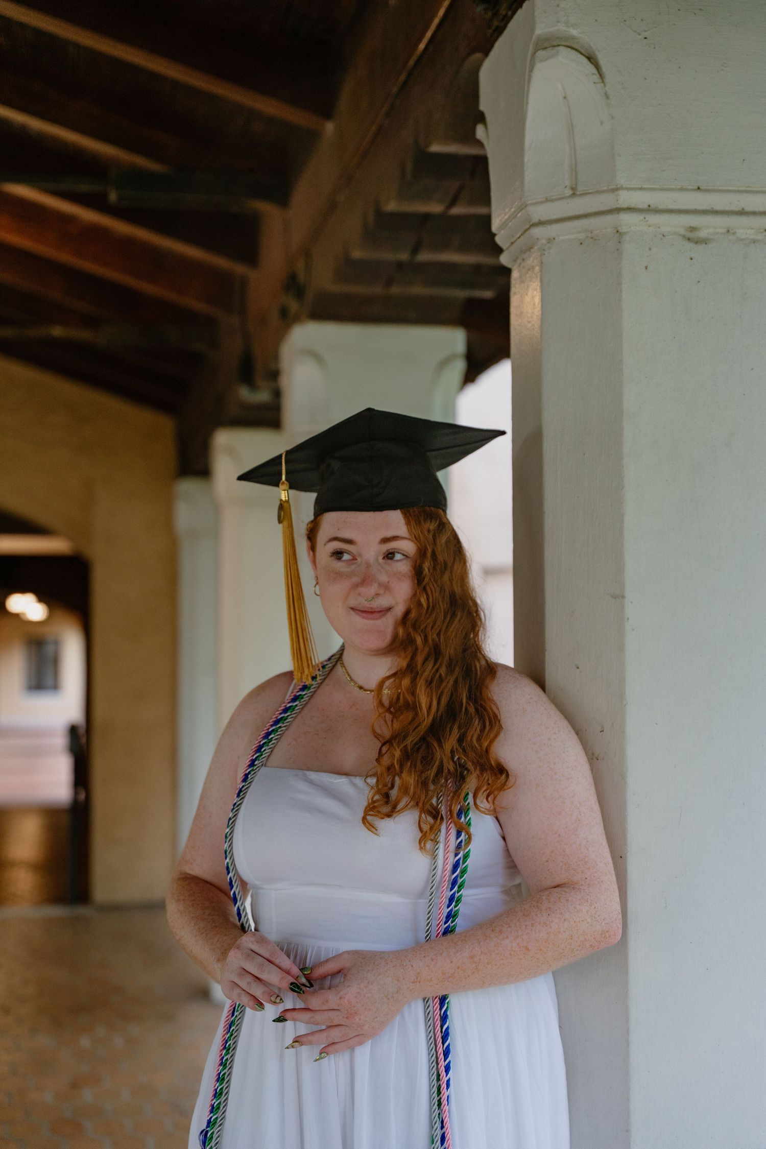Red-haired graduate in white dress and black mortarboard cap leans against white column for graduation portrait.