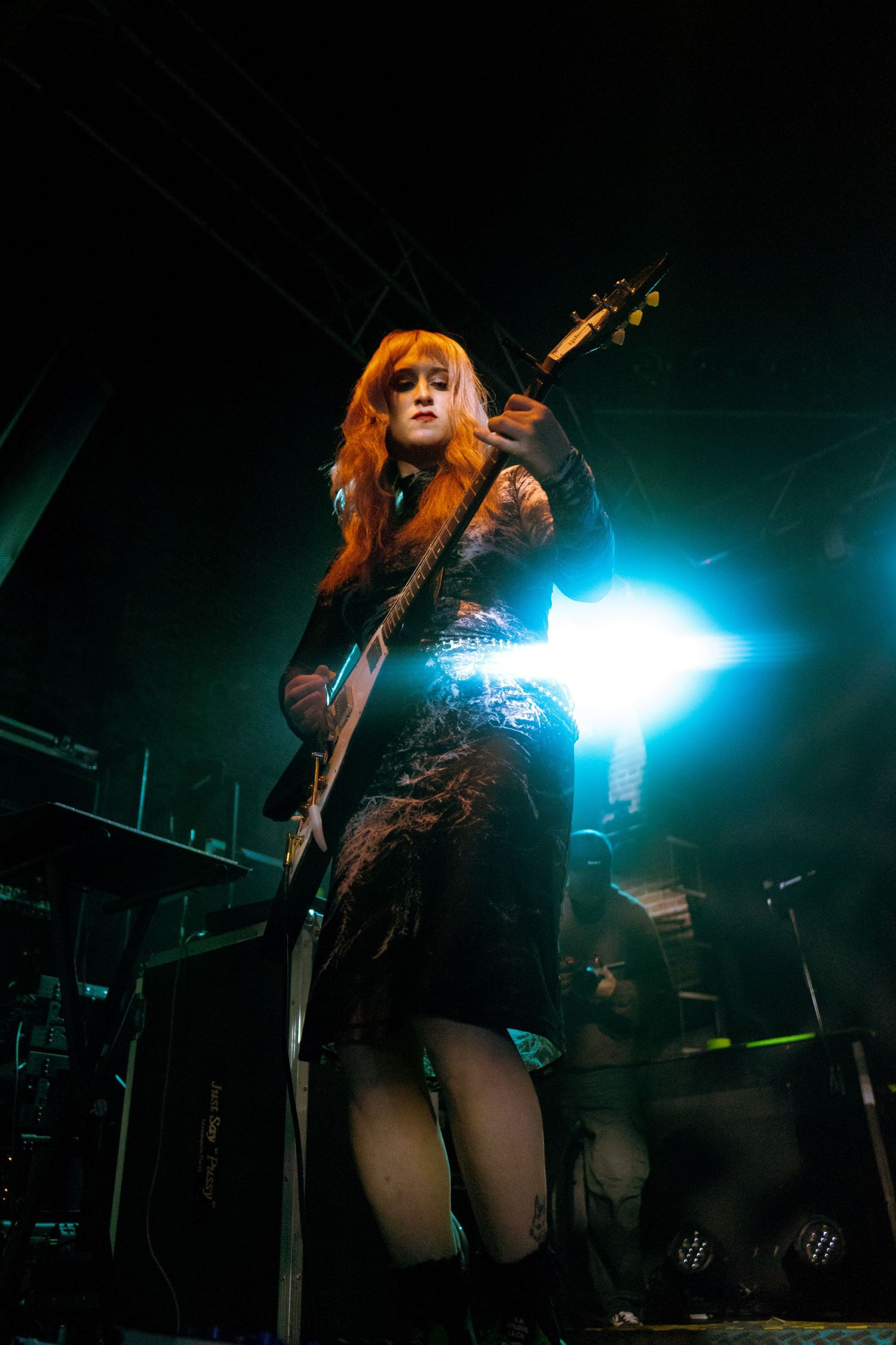 Red-haired female guitarist plays electric guitar on dark stage illuminated by bright blue backlight.