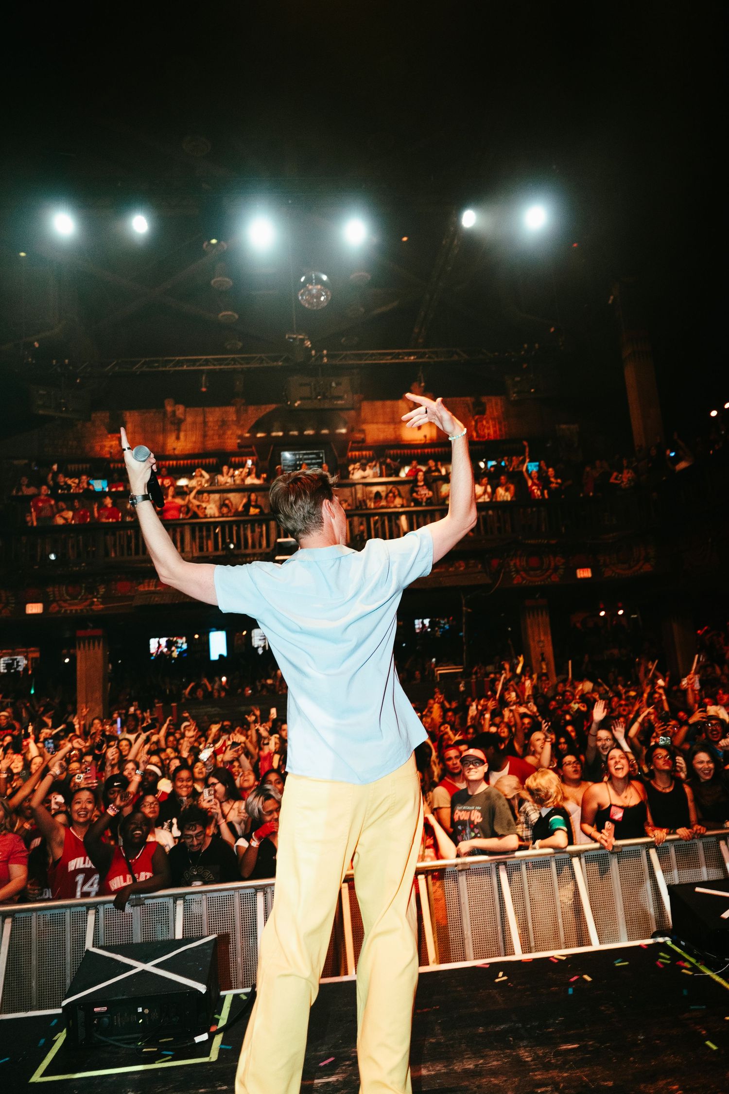 Performer in light blue shirt raises arms to energetic sold-out concert crowd from stage edge.