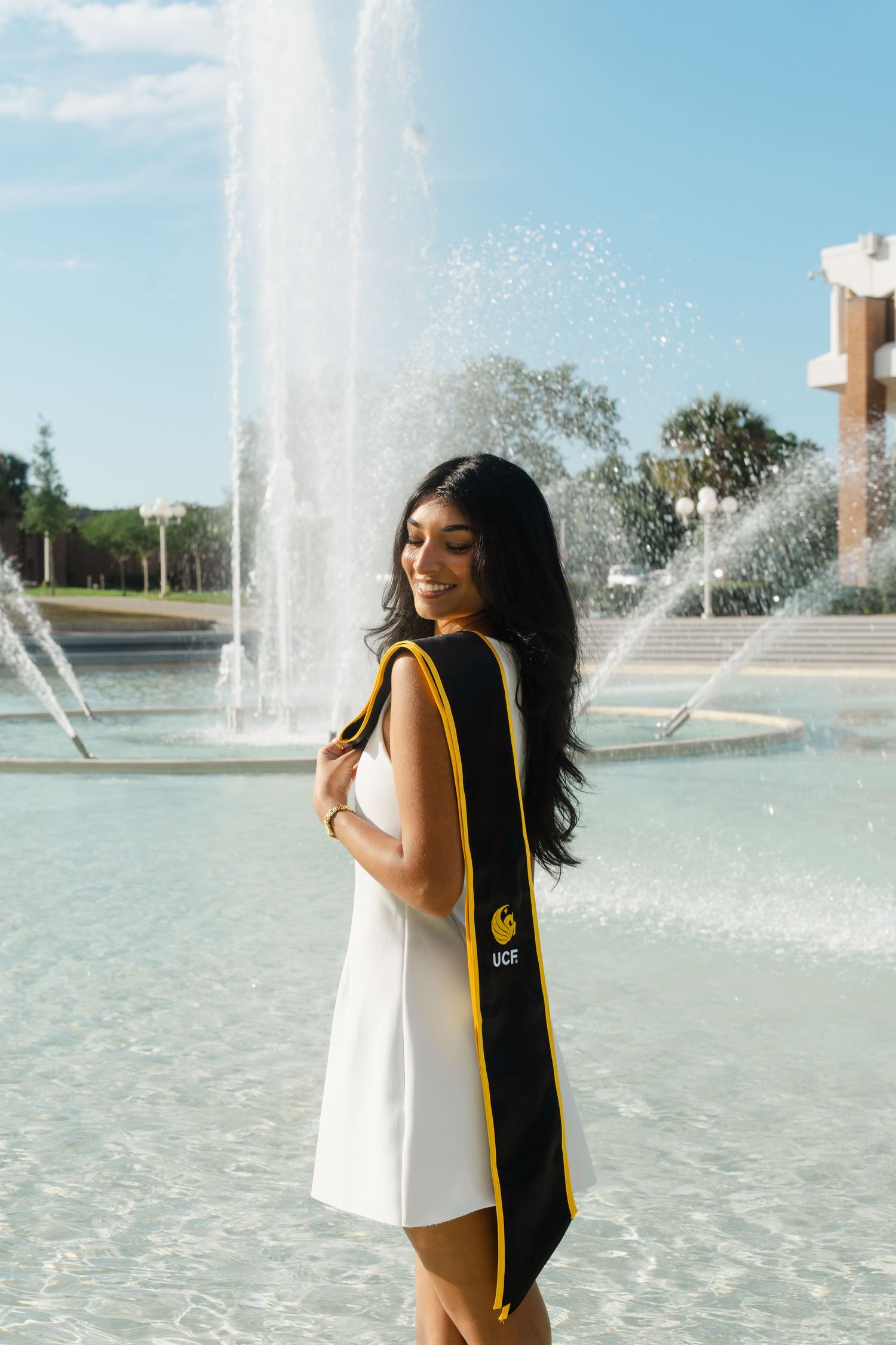 Young woman in white dress wearing UCF graduation stole smiles in front of a fountain on a sunny day.
