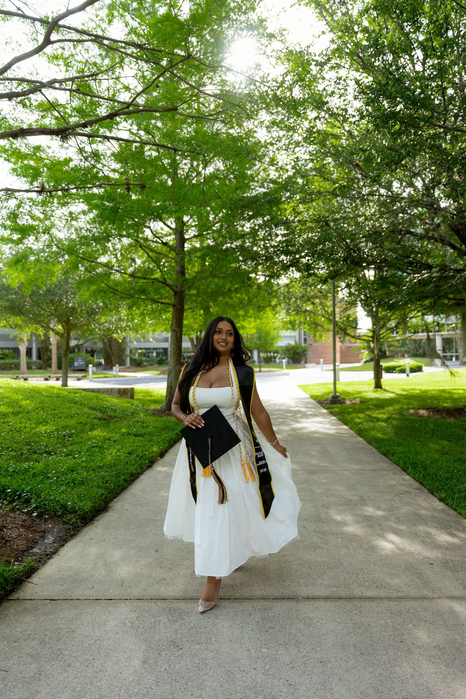 Smiling graduate in white dress and black cap and gown walks along tree-lined campus pathway.