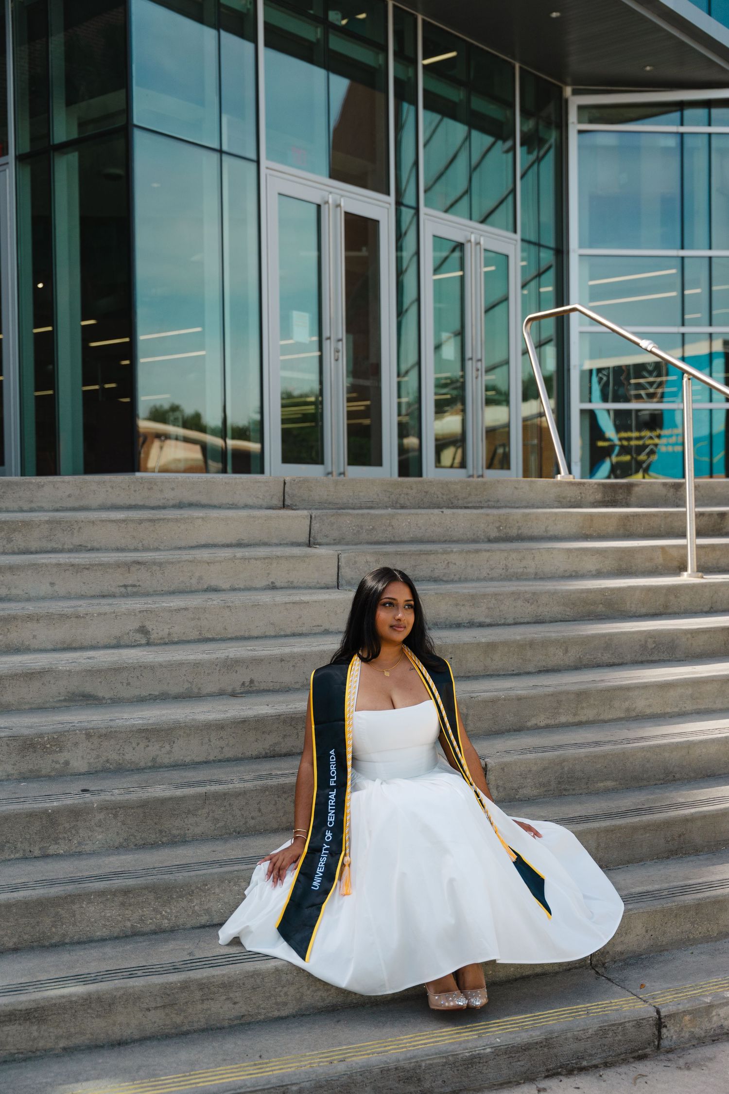 Graduate in white dress and black gold sash poses on university steps, twirling skirt in celebration.