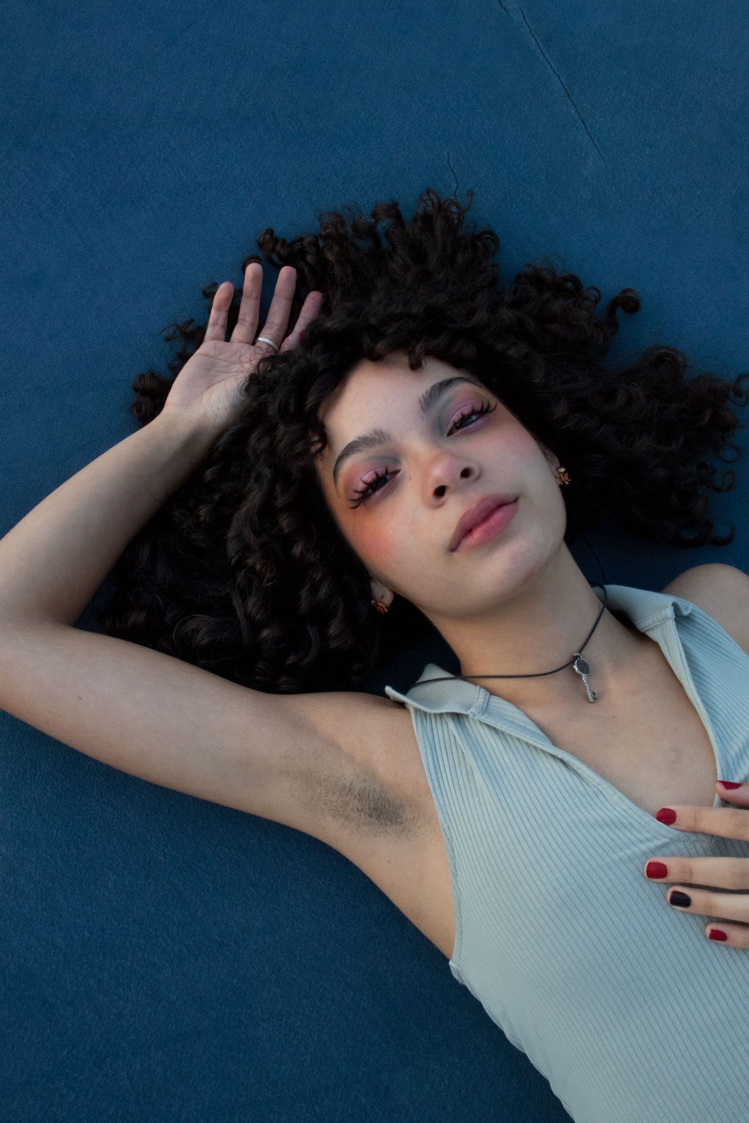 Young woman with curly dark hair lying on blue surface with colorful eye makeup.