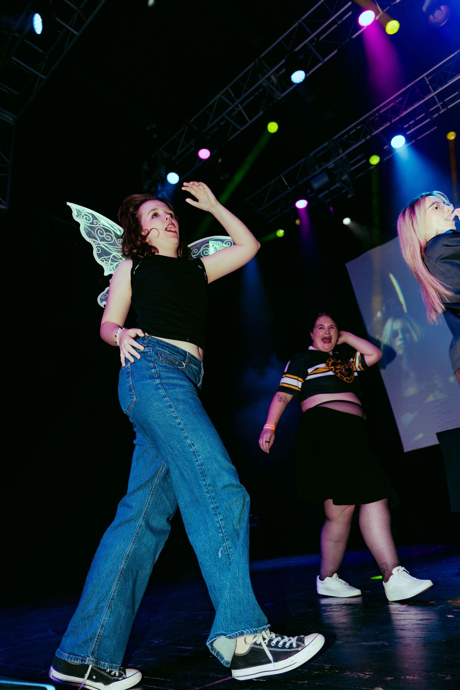 Two young women dancing on a lit stage, one wearing fairy wings, jeans, and black top under colorful concert lights.