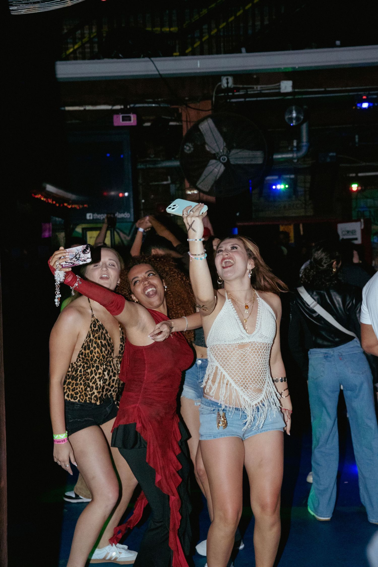 Three women dancing and taking selfies at a lively nightclub party with colorful lights.