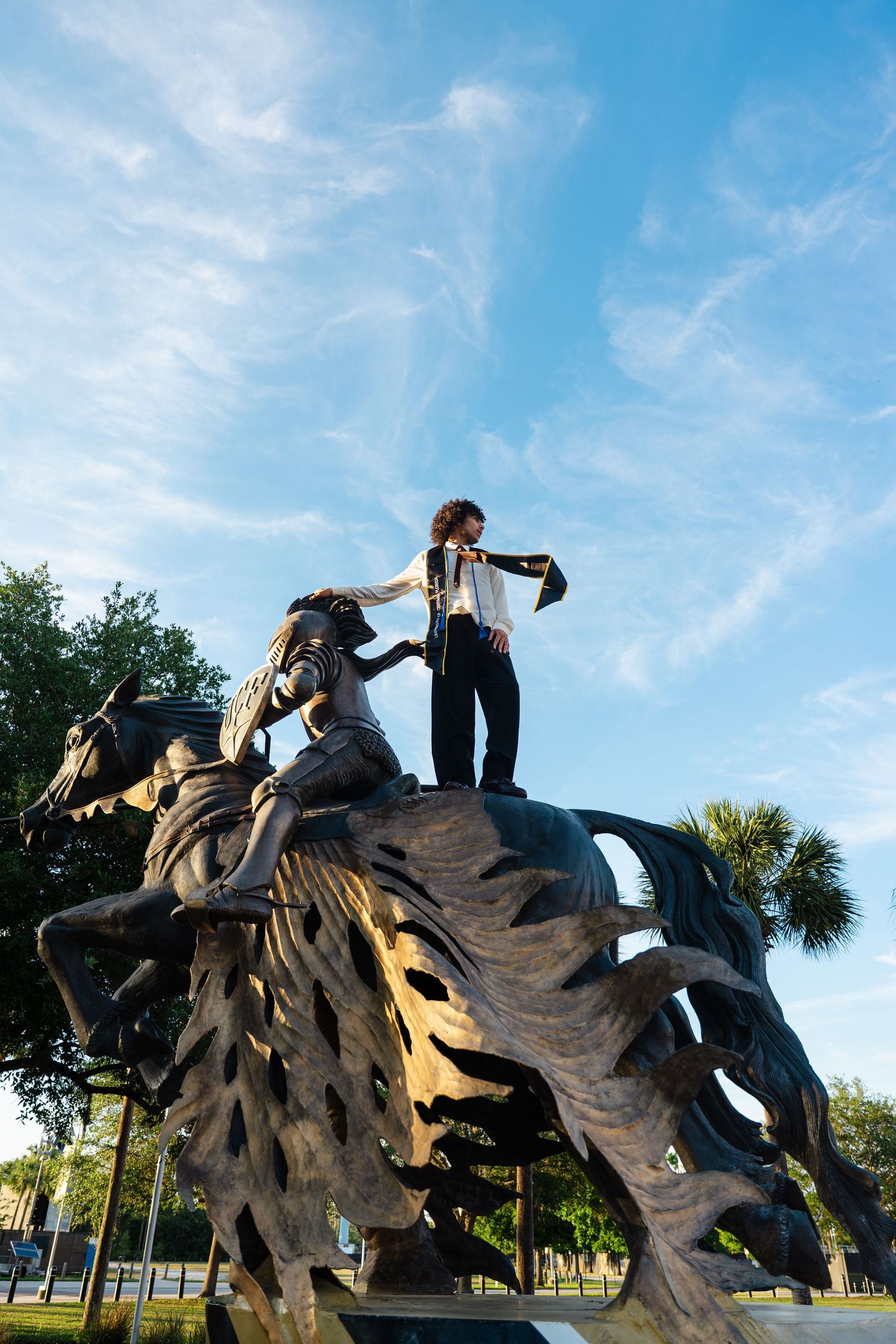 Person standing atop a large bronze sculpture outdoors, holding a scarf against a blue sky with wispy clouds.