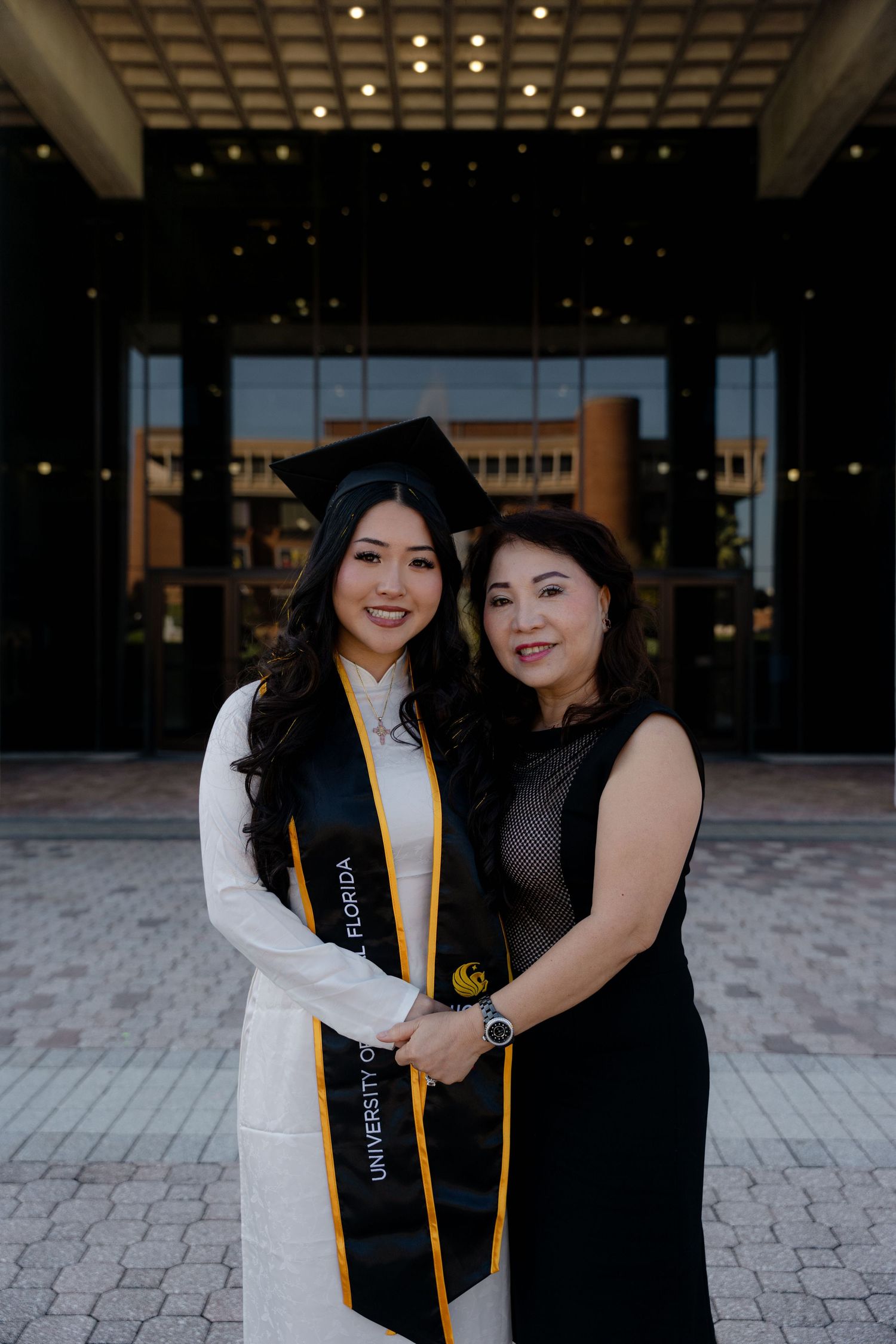 University of Florida graduate in white ao dai and gold stole poses with mother outside building.