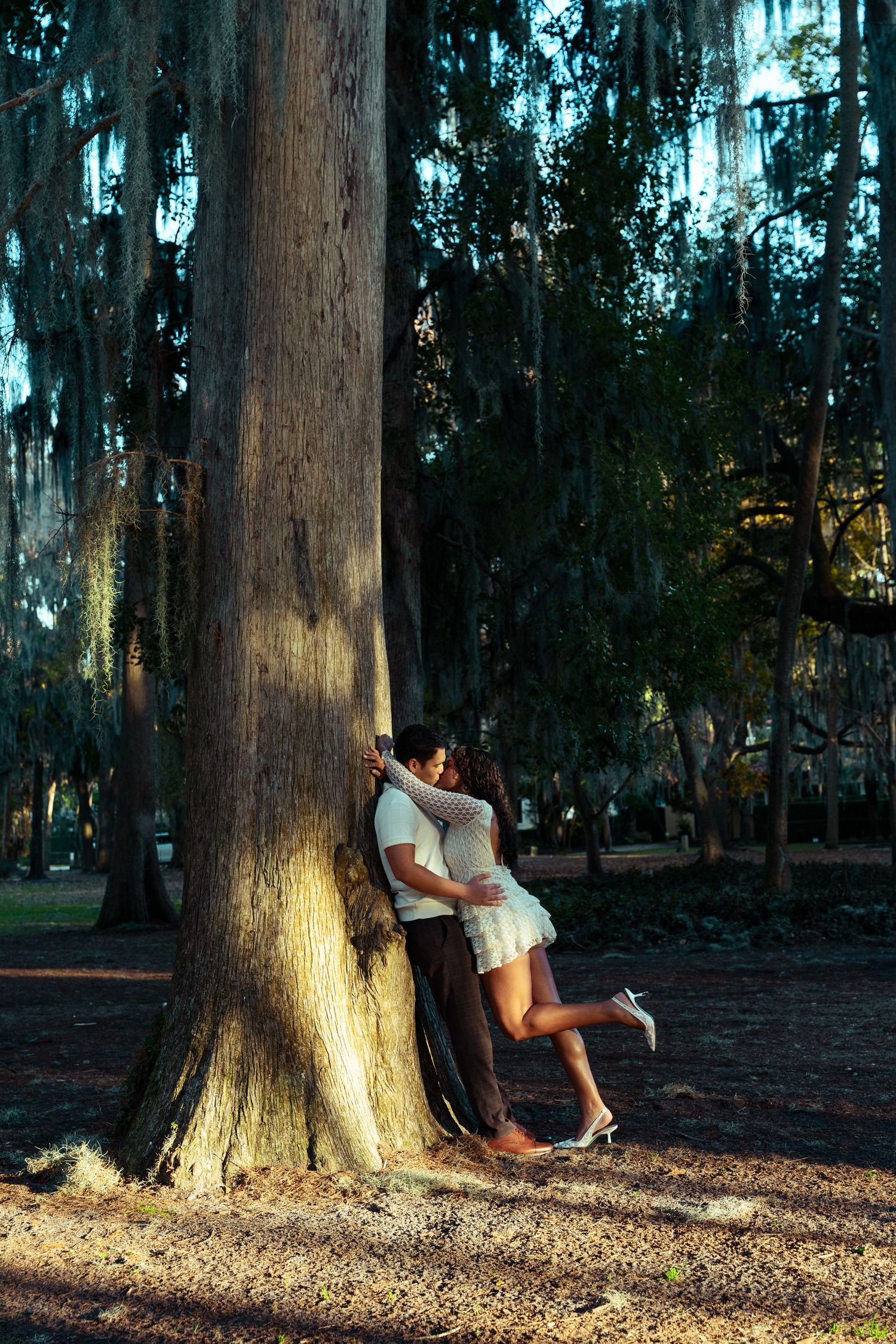 Couple sharing a romantic embrace against a large tree trunk in a sunlit moss-draped park.