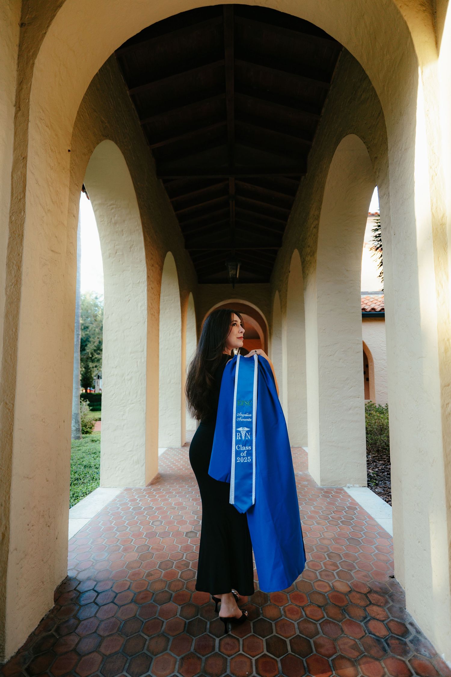 Graduate in black outfit draped in blue stole poses under arched corridor in golden light.