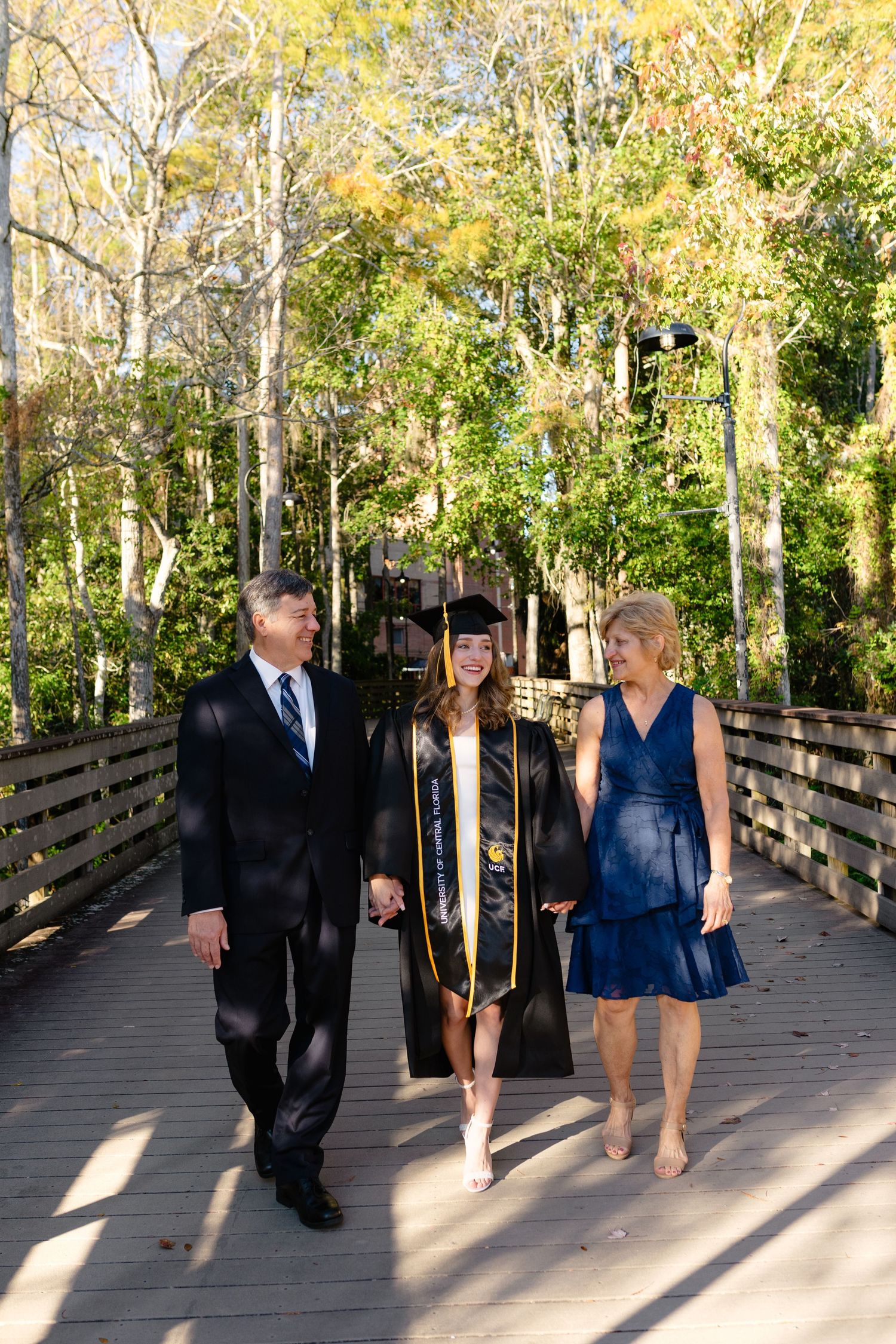 Graduate in cap and gown walking with proud parents on a tree-lined boardwalk.