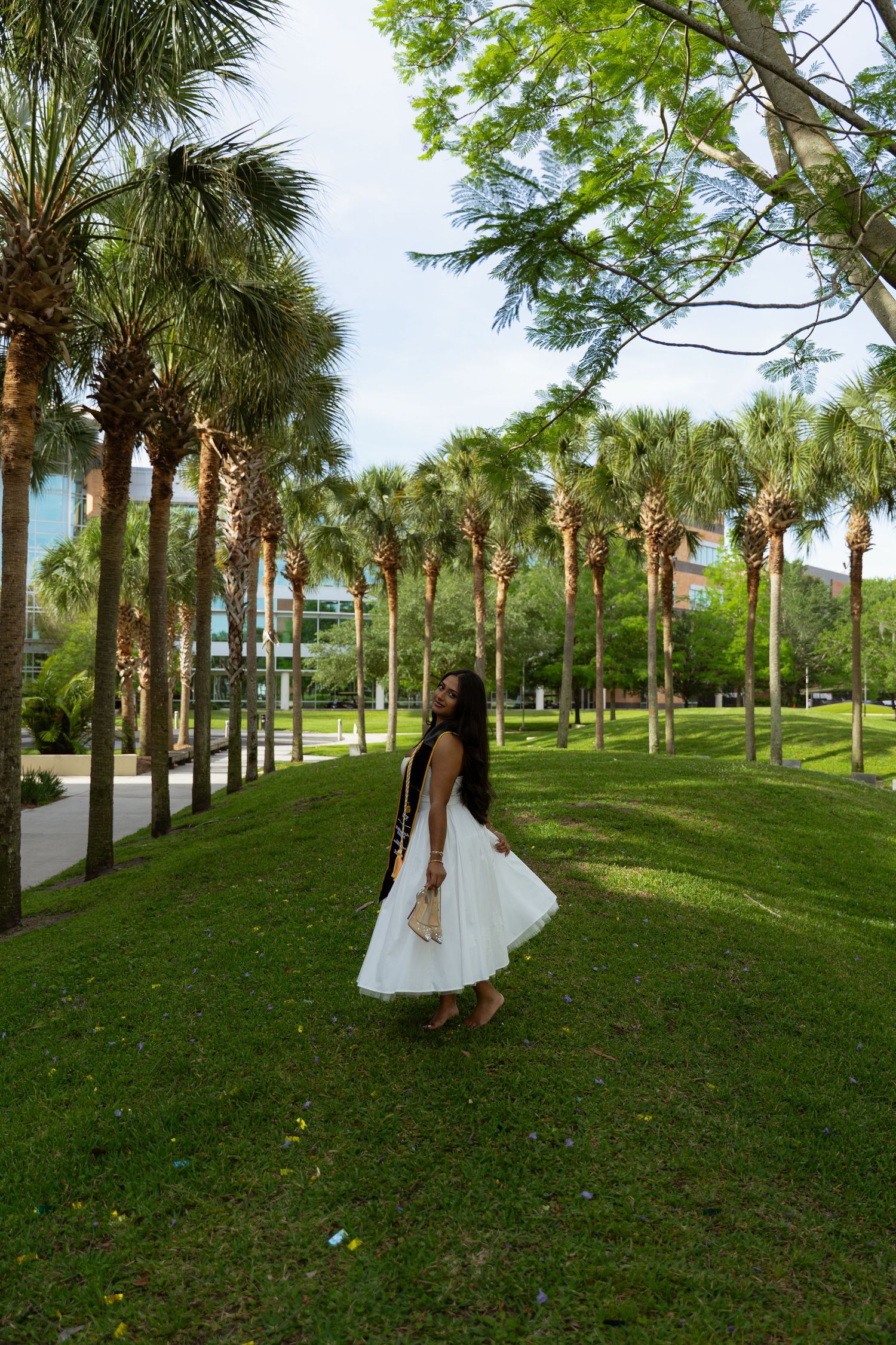 Woman in white dress standing among palm trees on lush green grass in a tropical park setting.