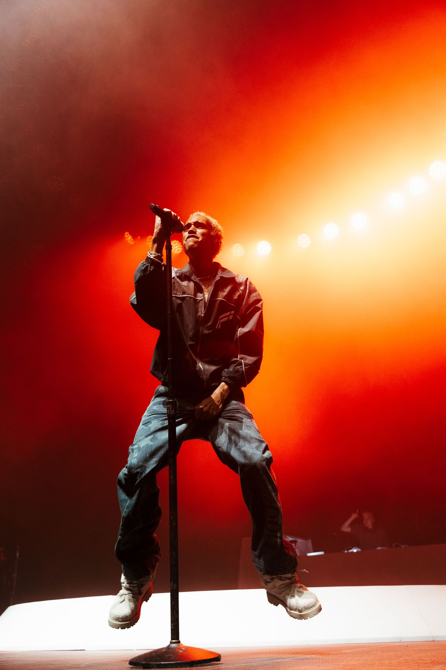 Performer jumps mid-air on stage holding microphone, bathed in dramatic red and white concert lighting.