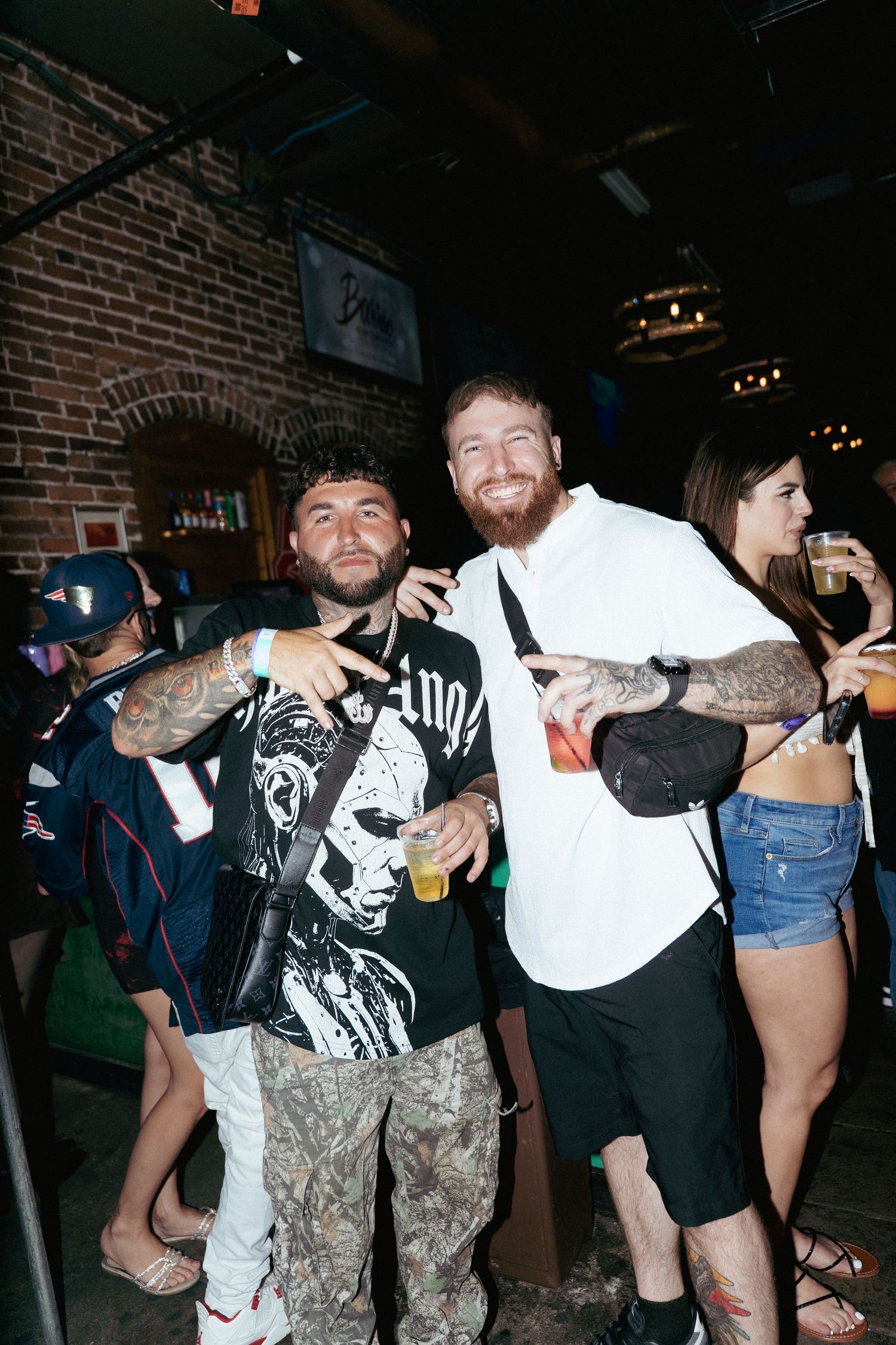 Two tattooed men pose together at a dimly lit bar, one in a graphic tee and camo pants, the other in a white shirt.
