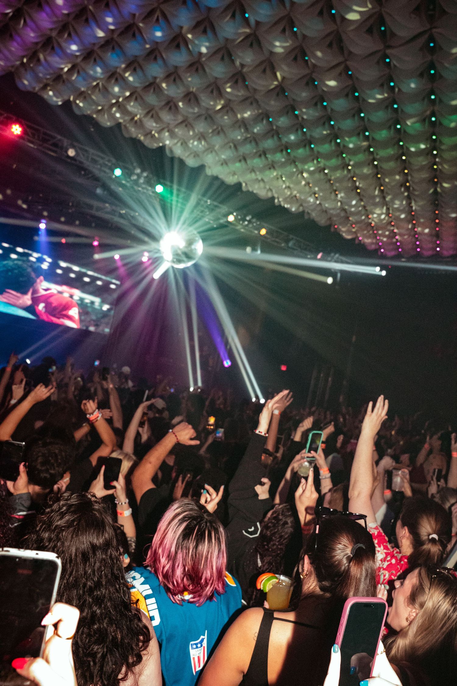 Crowd of concert-goers with hands raised under colorful disco ball lights at a packed indoor music event.