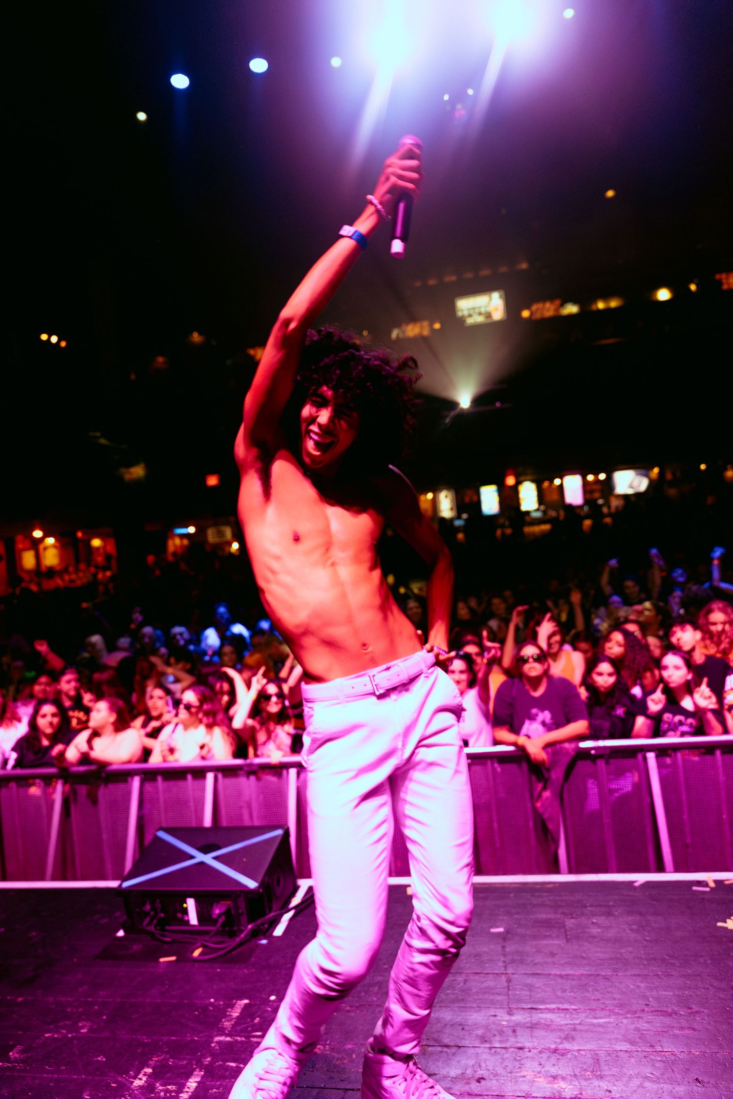 Shirtless male performer holds microphone high on stage under pink lights before a large concert crowd.