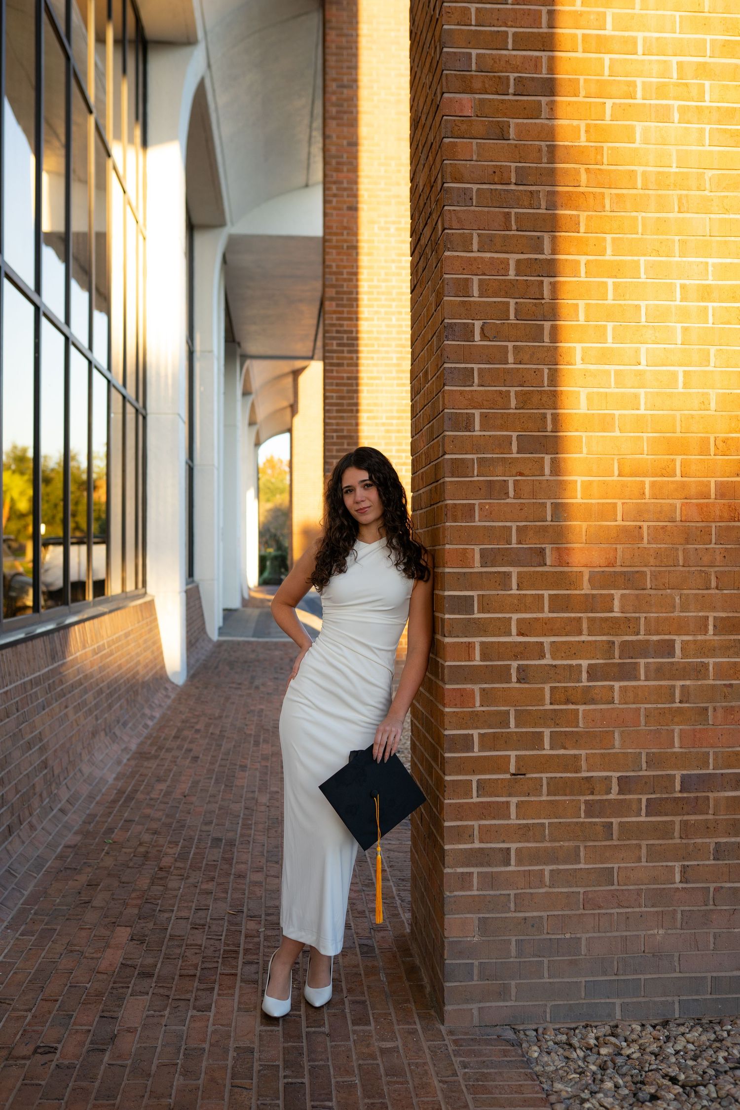 UCF graduate in white dress holds cap while leaning against brick wall in golden sunset light.