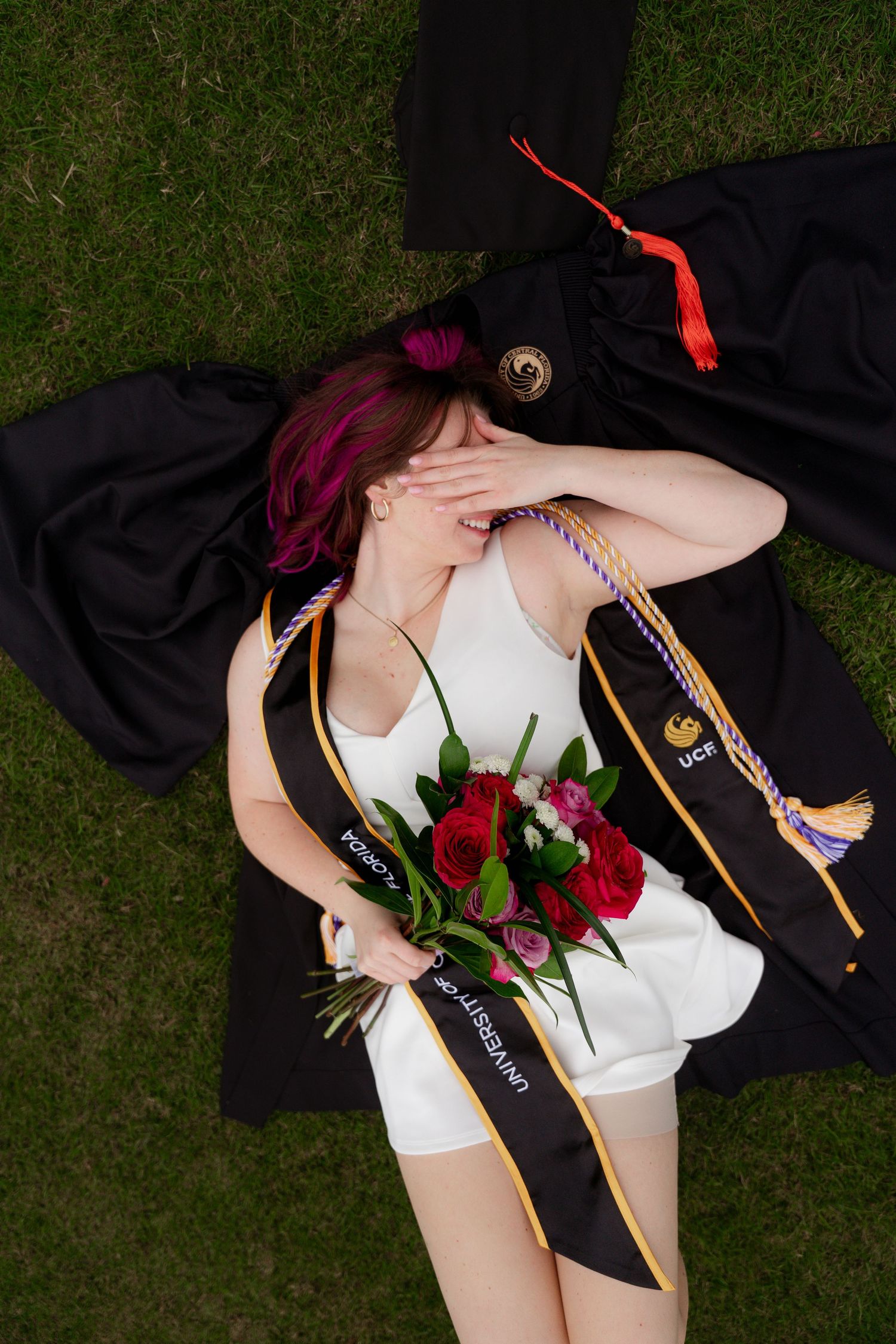 Female graduate lies on grass with UCF sash, bouquet of red roses, and black graduation gown.