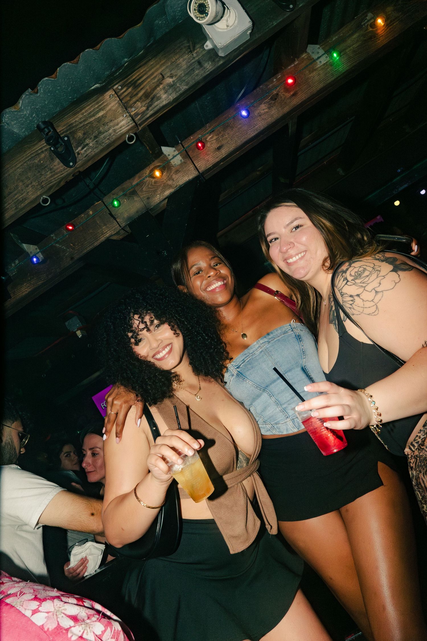 Three women smiling and holding drinks at a dimly lit nightclub with colorful string lights.