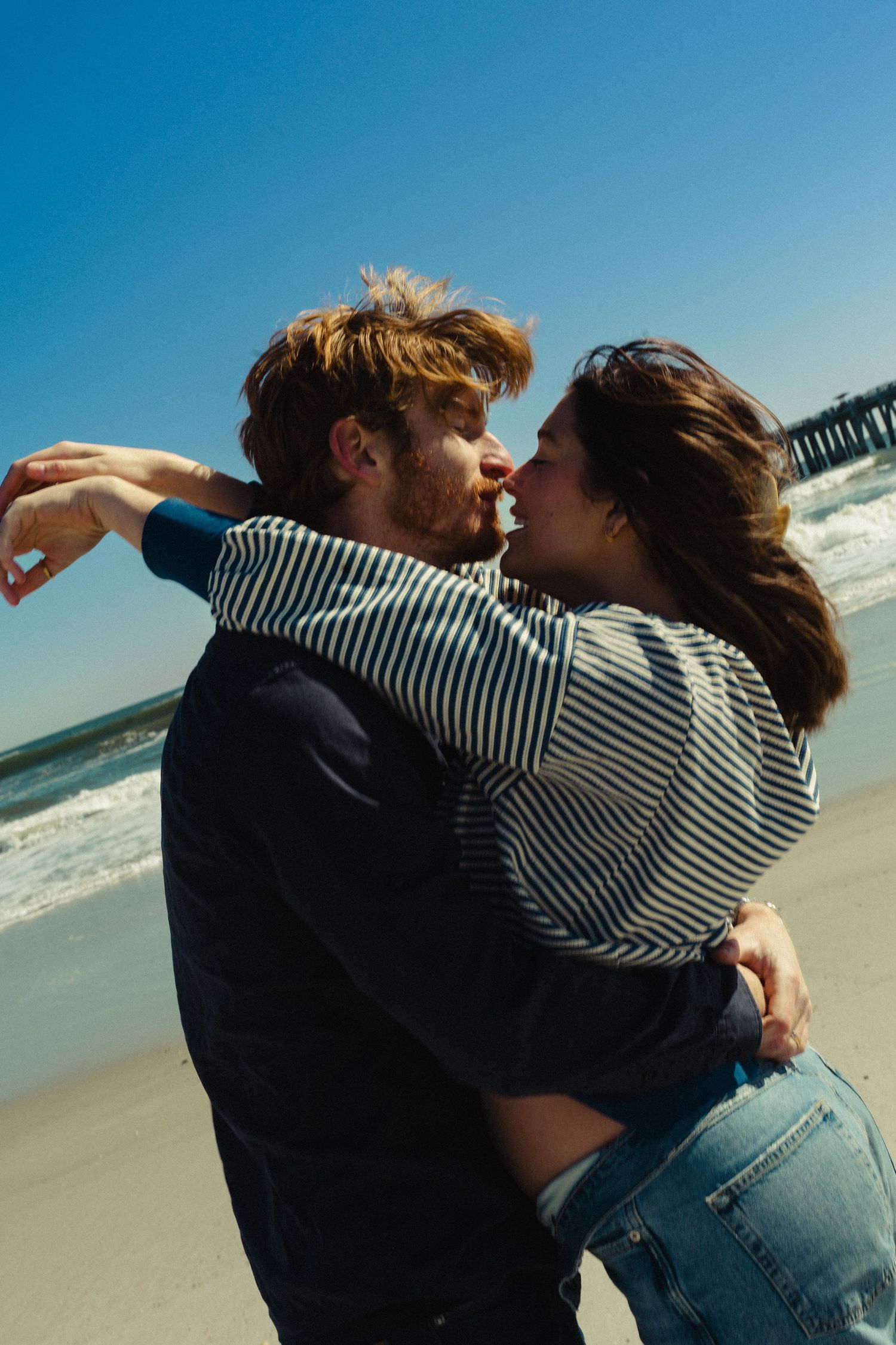 A couple embracing and kissing on a sunny beach with ocean waves and a pier in the background.