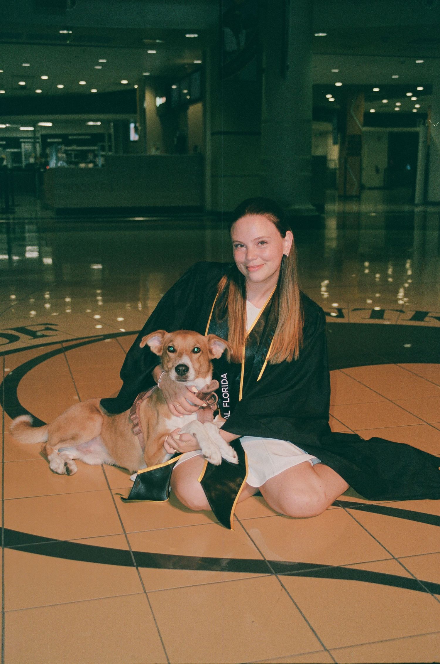 Young woman in graduation gown sitting on tiled floor with a brown and white dog in an indoor venue.