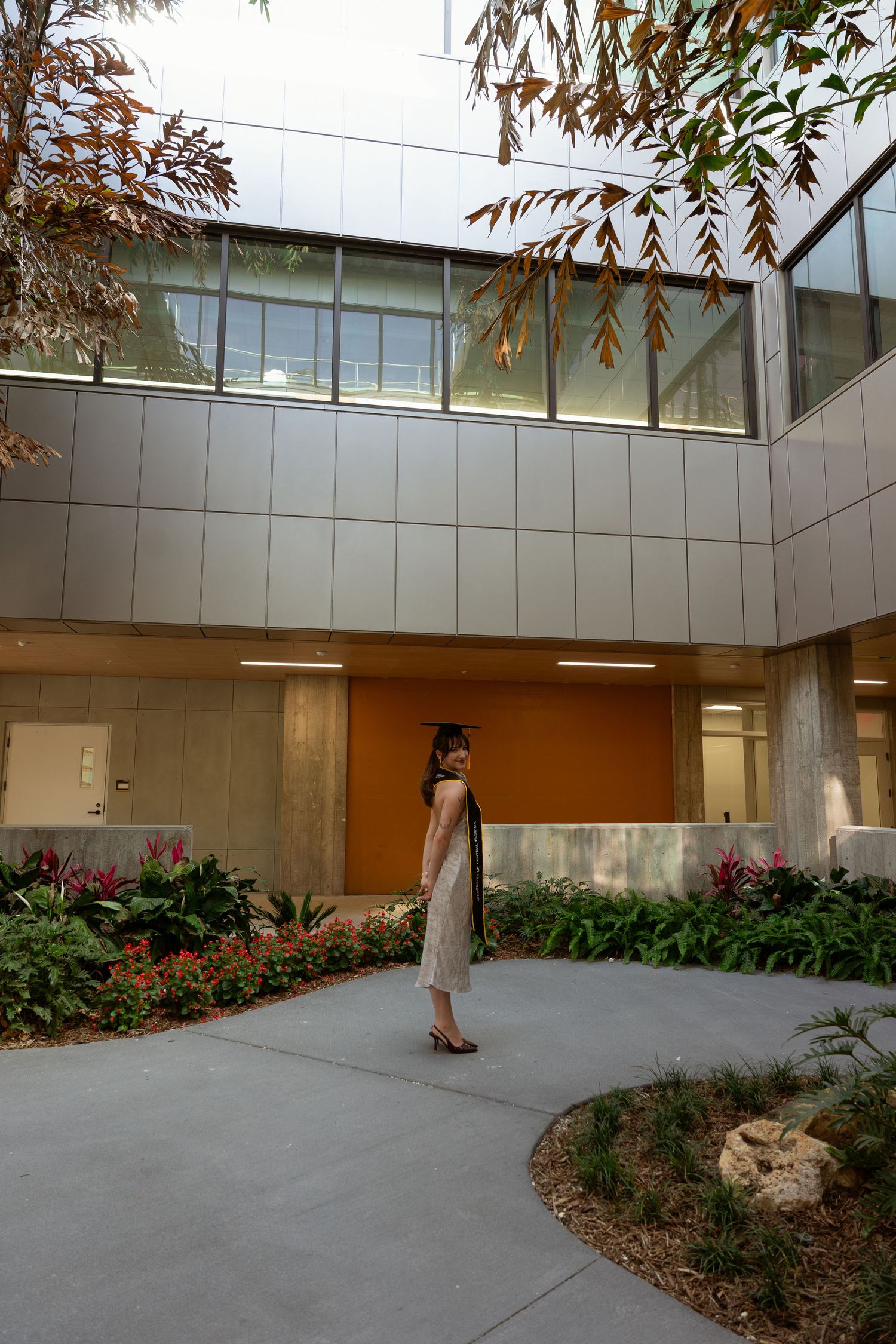 Woman in white dress standing in modern building courtyard with lush tropical plants and glass facade.