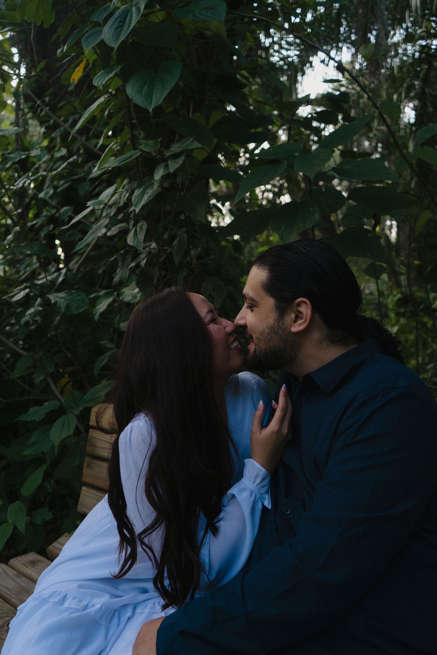 Romantic couple sharing a kiss while sitting outdoors surrounded by lush green tropical foliage.