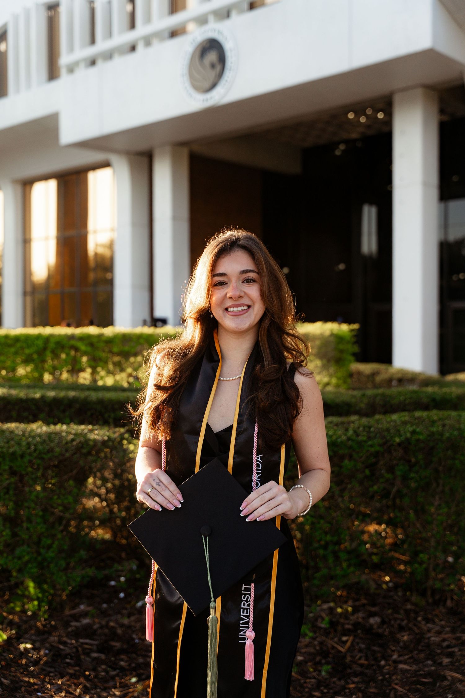 Smiling female graduate holds black cap with colorful tassels outside university building.
