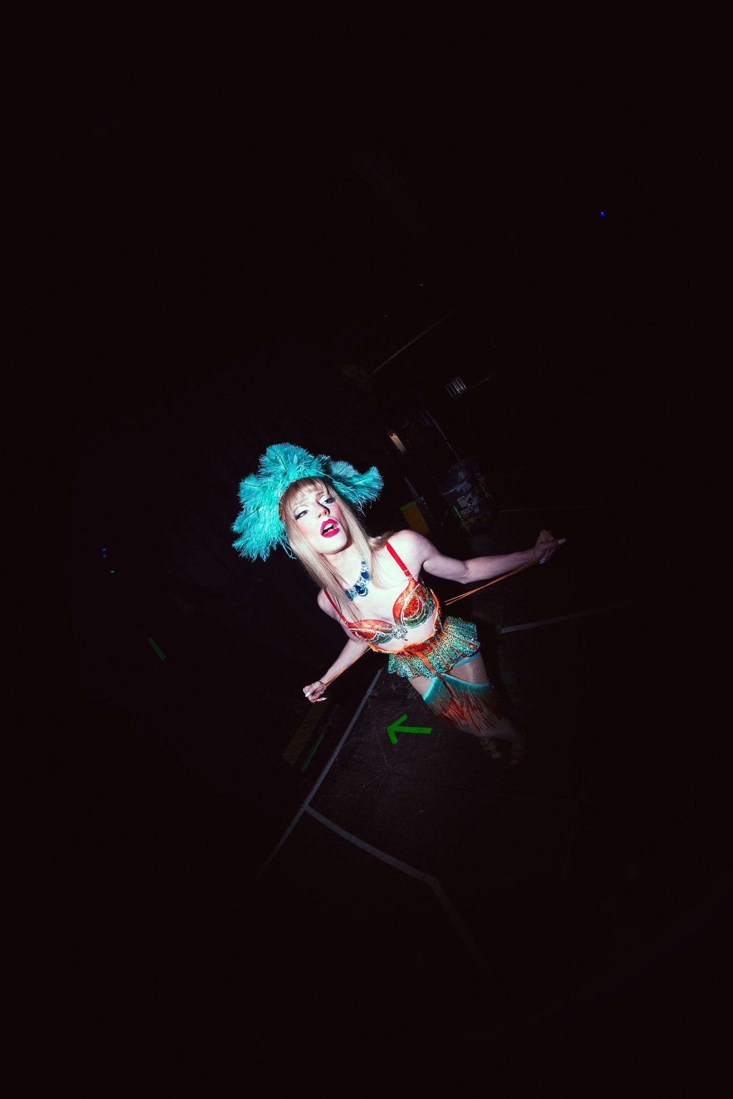 Female performer in colorful costume with turquoise feather headpiece dancing on dark stage.