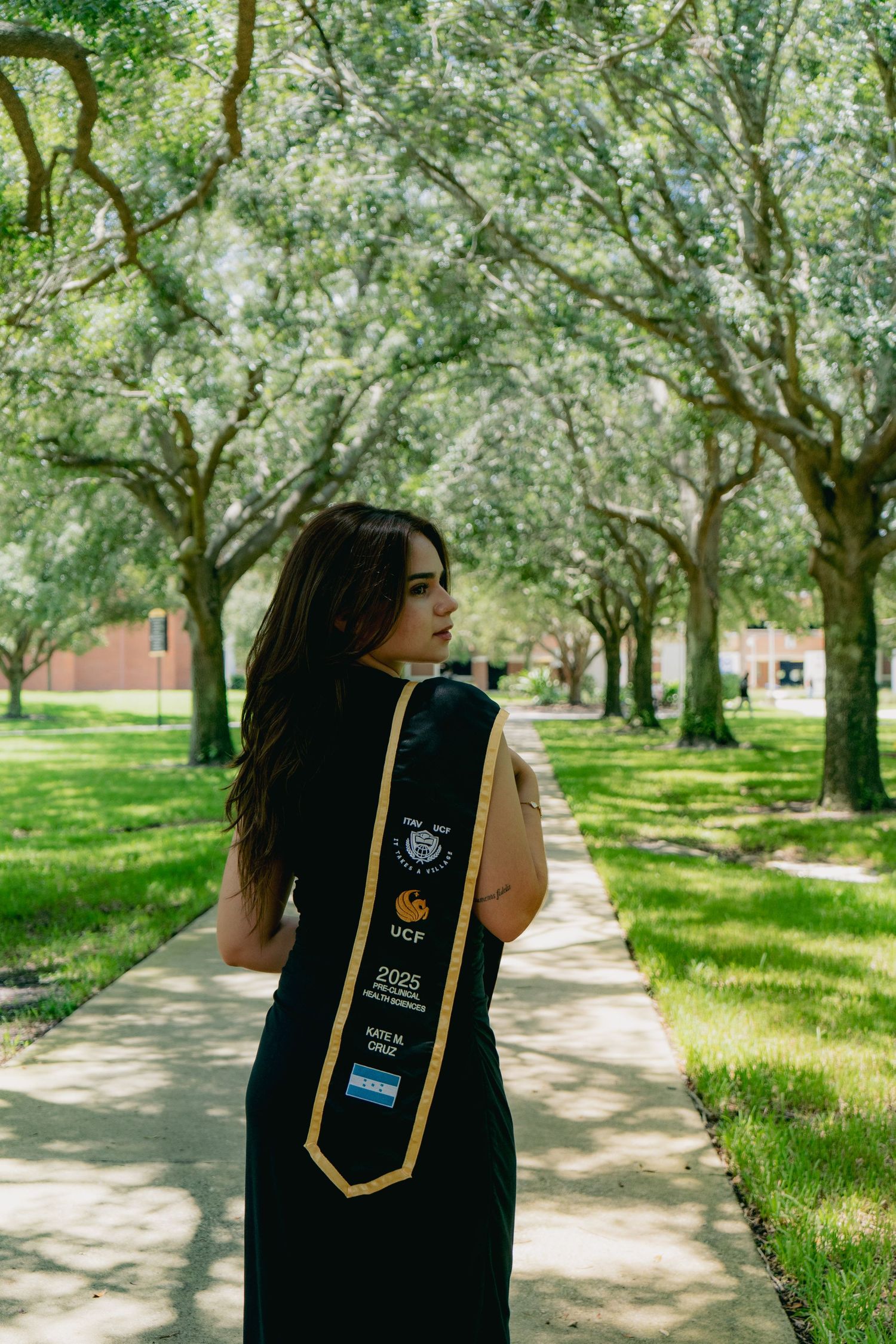 UCF 2025 graduate in black gown walks along a sunlit tree-lined campus pathway displaying graduation stole.