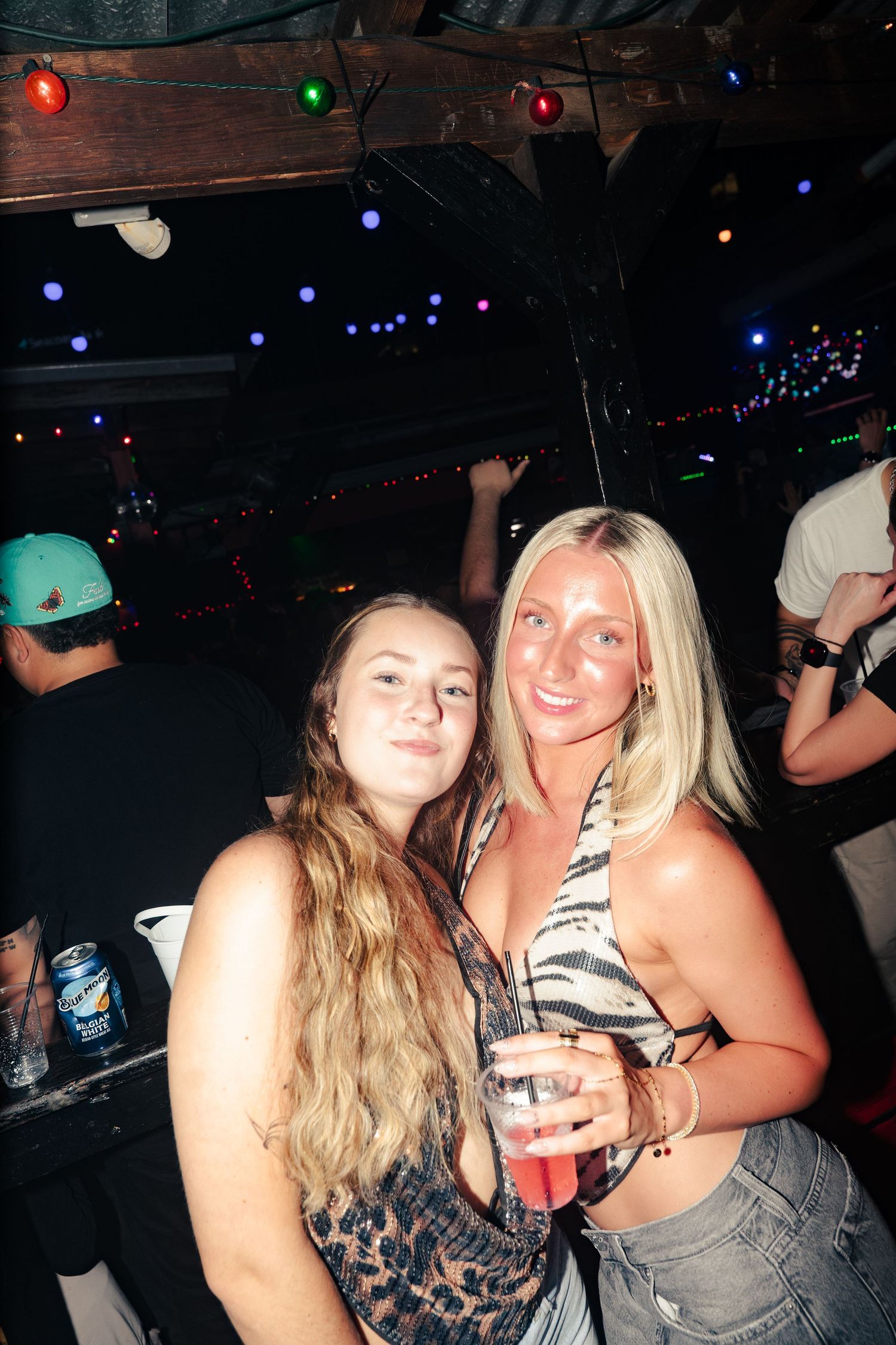 Two young women smiling together at a nightclub, one with brown hair and one with blonde hair holding a drink.