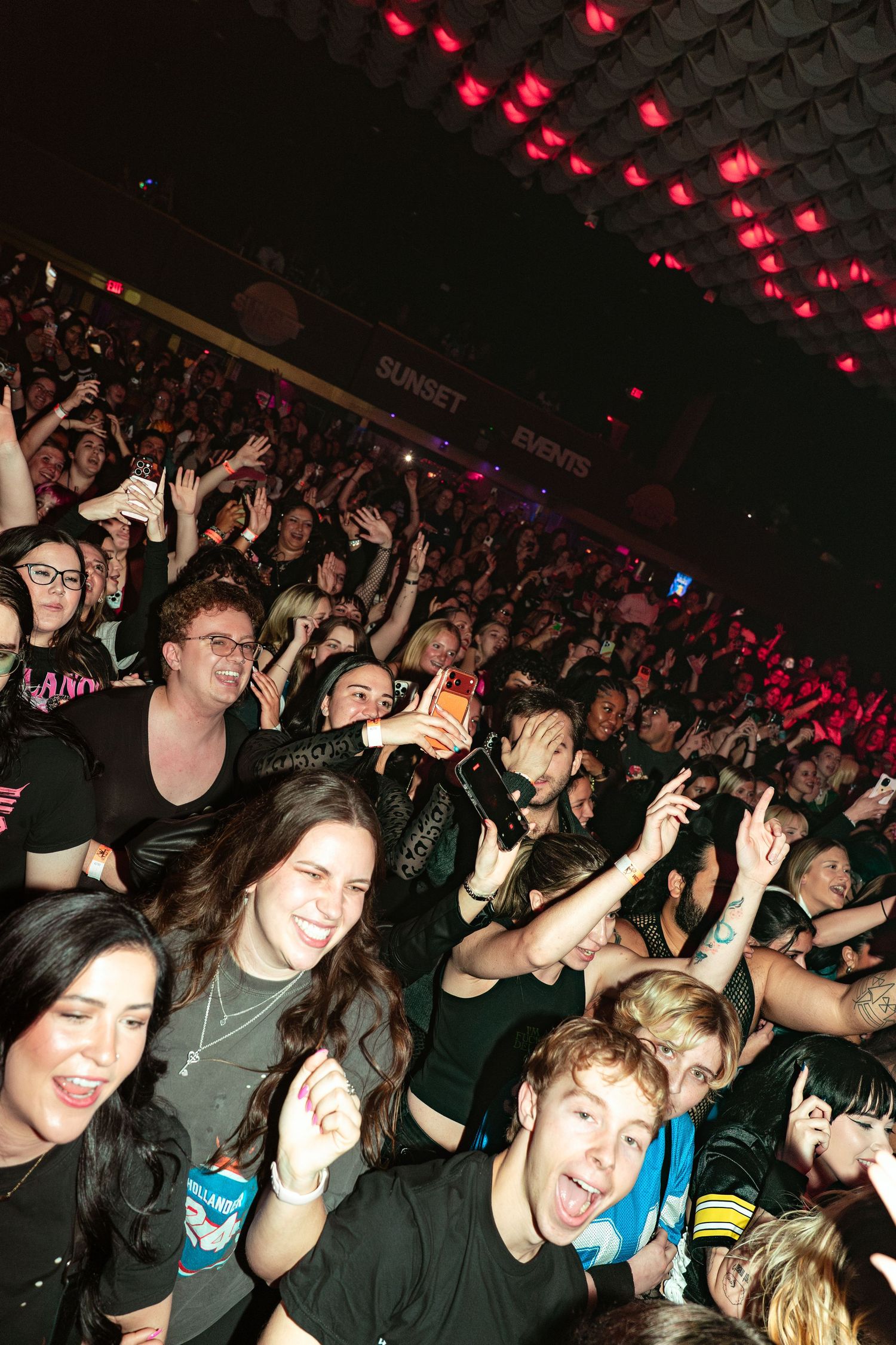 Excited concert crowd with hands raised under red lights at a packed indoor music venue.