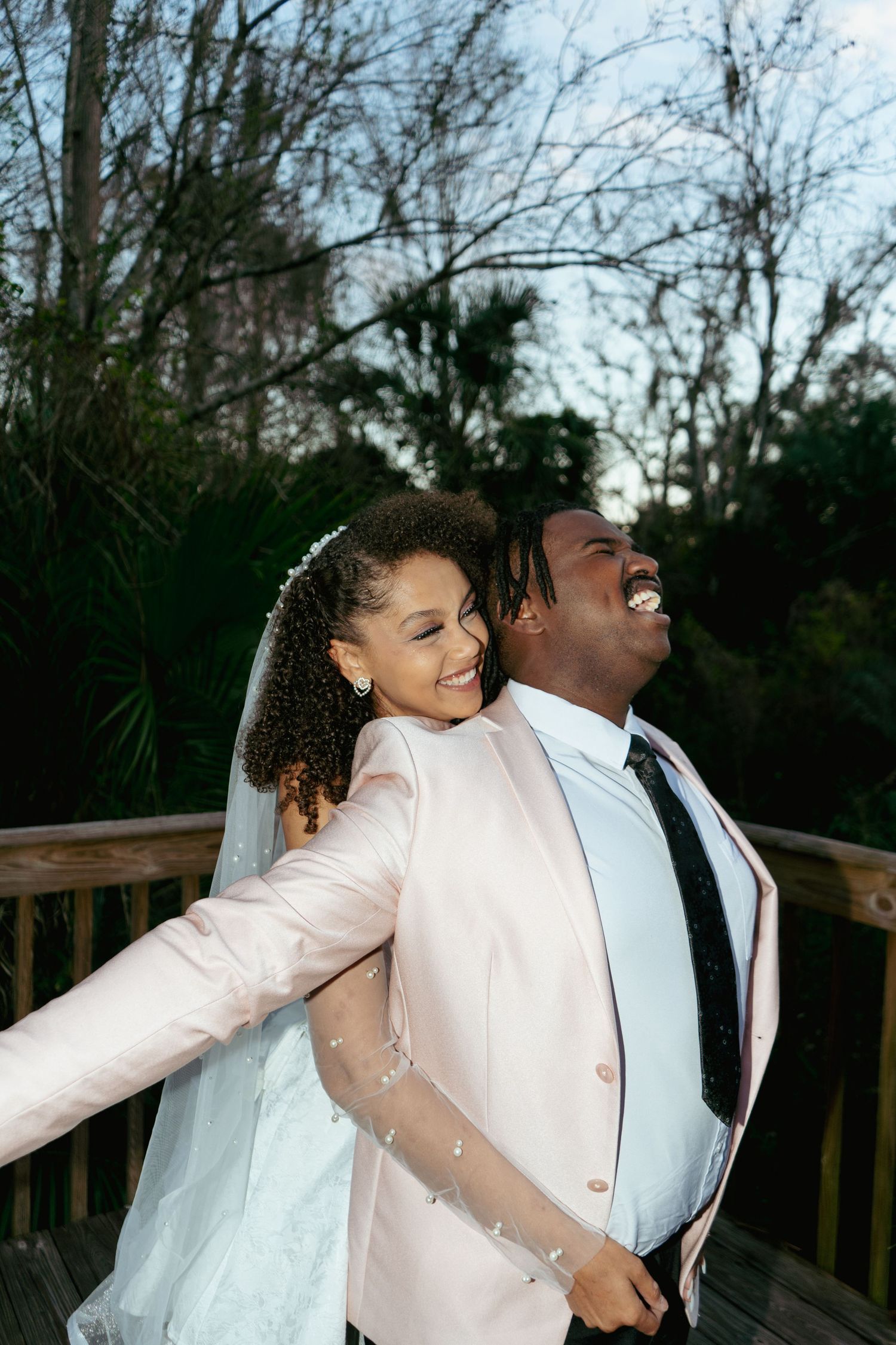 Happy bride and groom embrace on an outdoor wooden deck surrounded by trees at dusk.