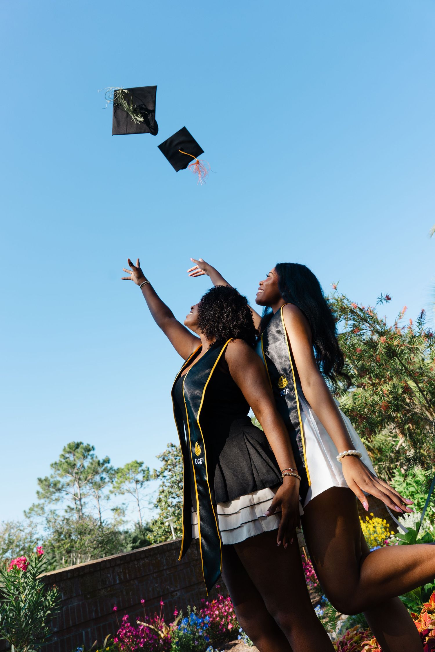 Two young women celebrating graduation by tossing a cap into the blue sky outdoors.