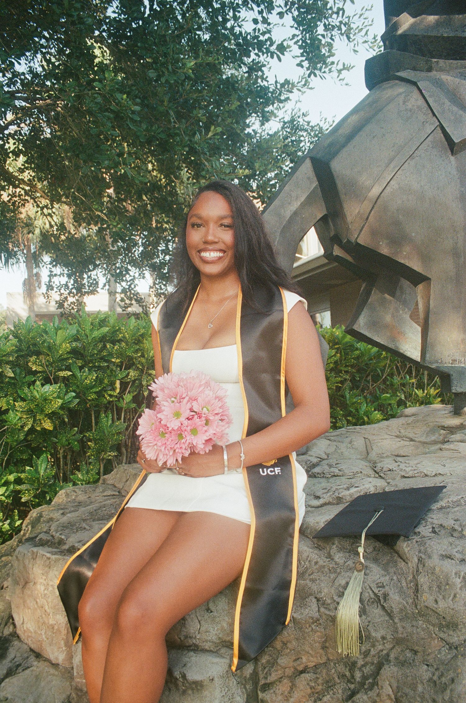 Smiling female UCF graduate holds pink flowers while sitting on rocks outdoors.