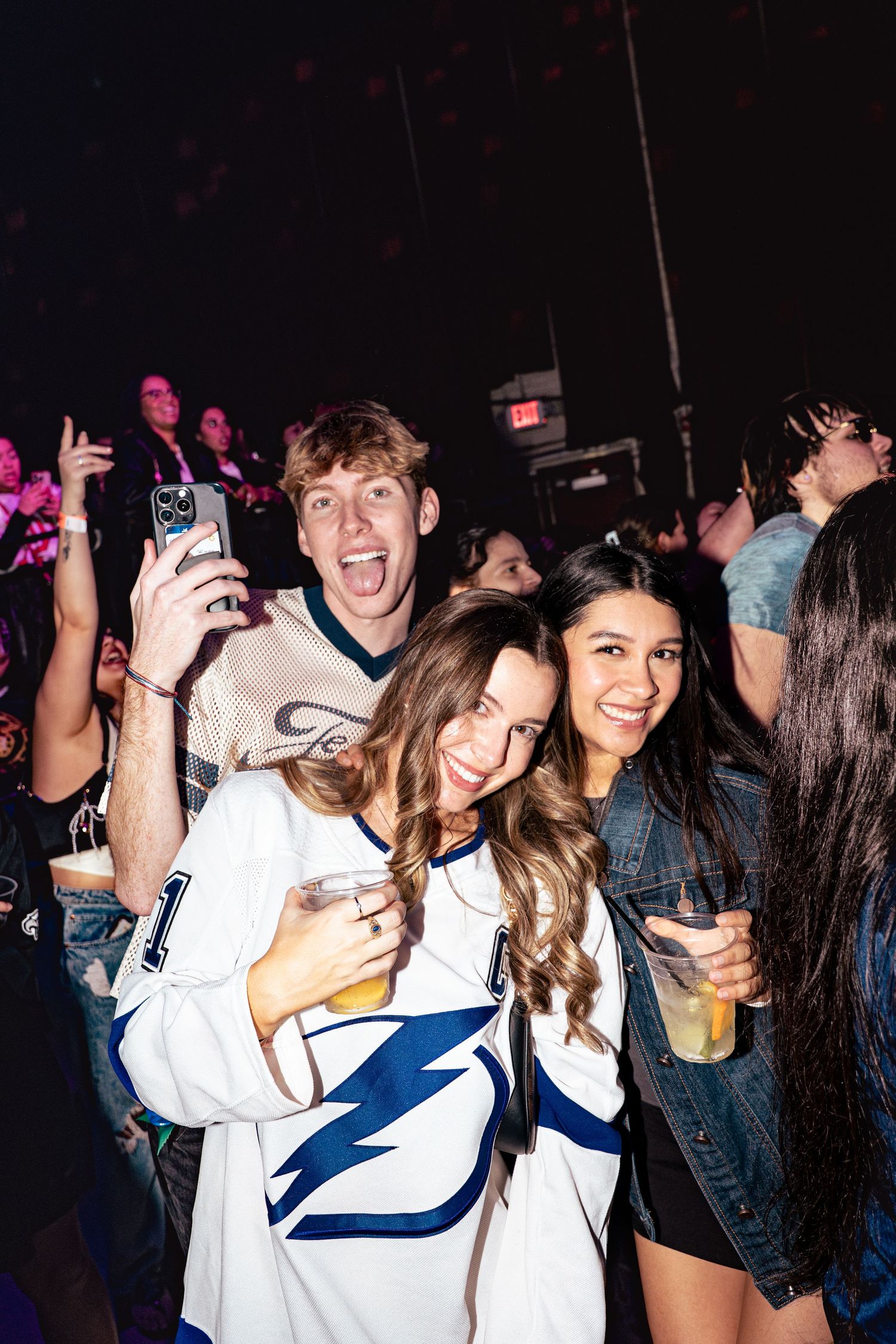 Three friends pose at a lively concert, one wearing a Tampa Bay Lightning jersey, holding drinks and smiling.