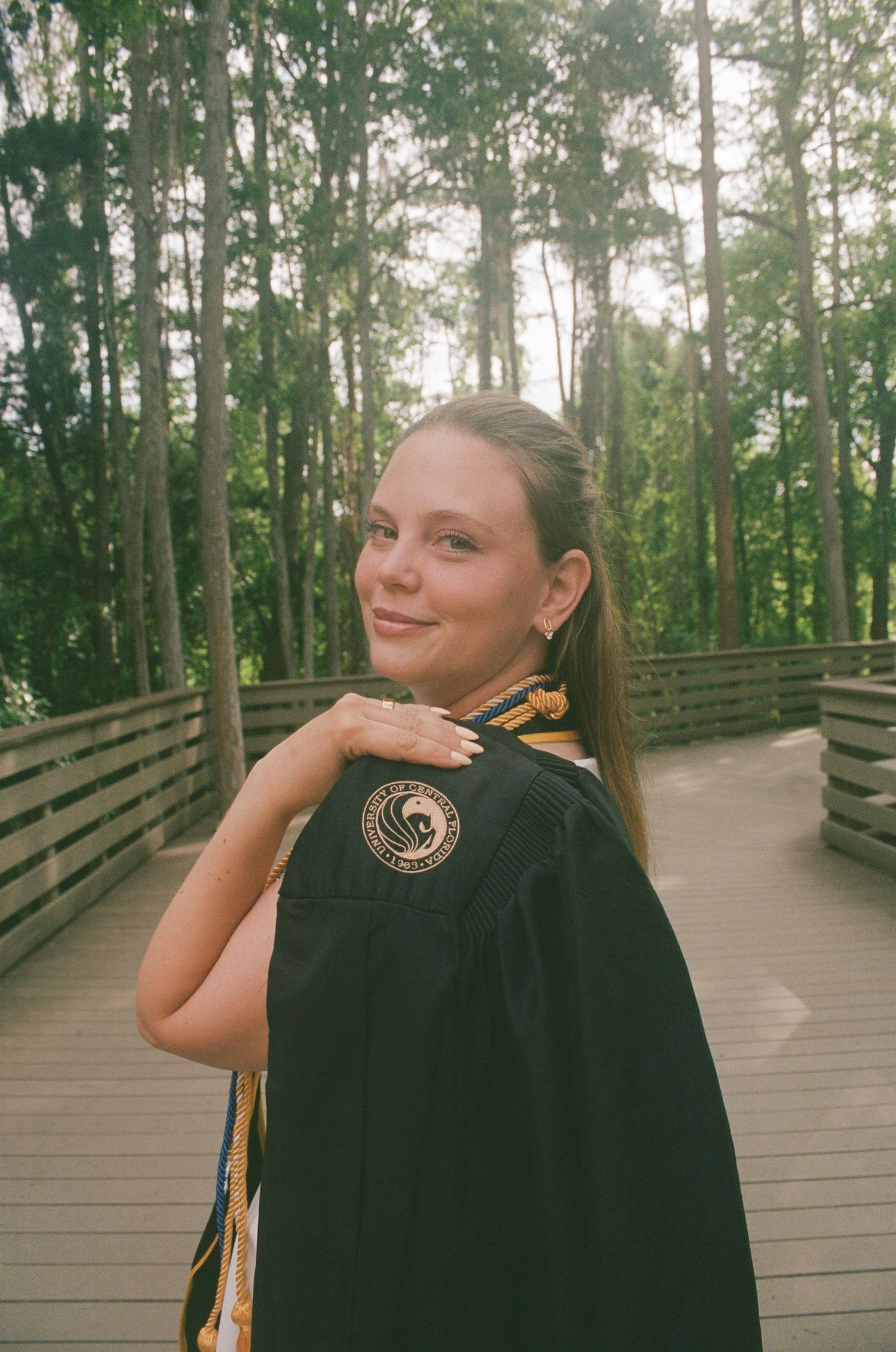 Young woman in black graduation gown with gold tassel smiles on a wooden bridge surrounded by tall trees.