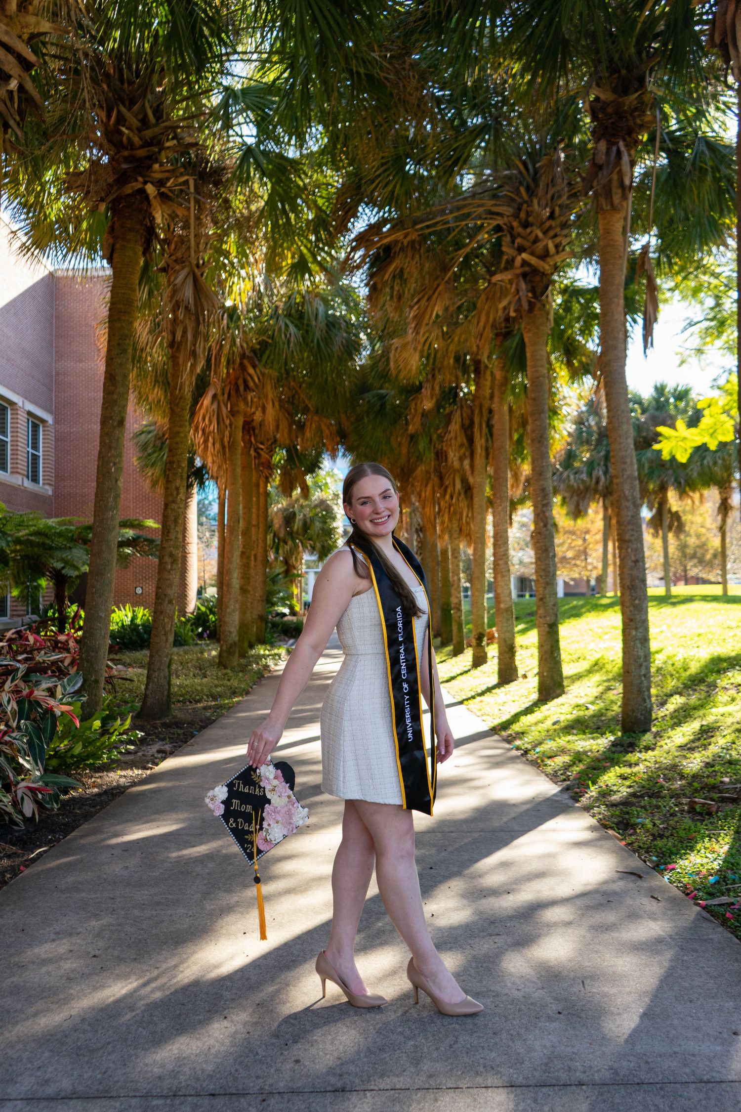 Smiling graduate in white dress and black sash walking along a palm tree-lined campus pathway.