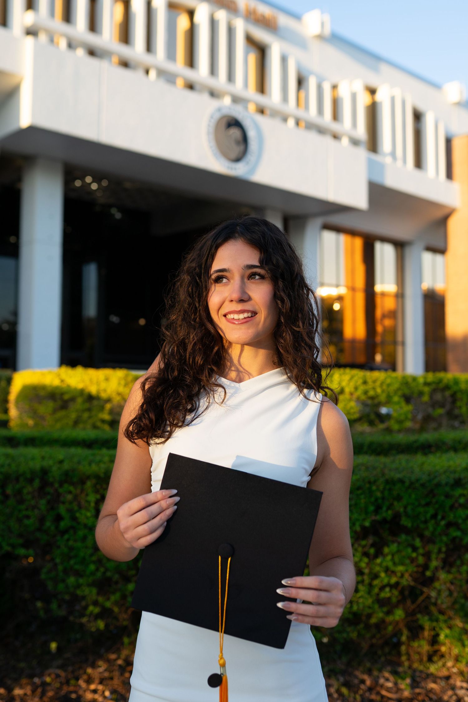 Smiling UCF graduate holds black cap with gold tassel outside Millican Hall at golden hour.