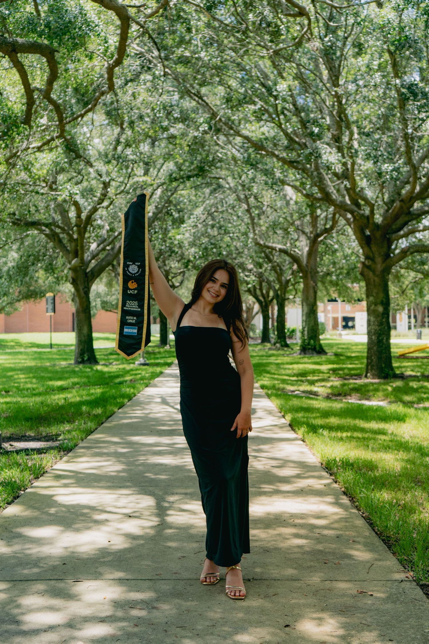 UCF graduate in black maxi dress holding graduation stole on tree-lined campus walkway outdoors.
