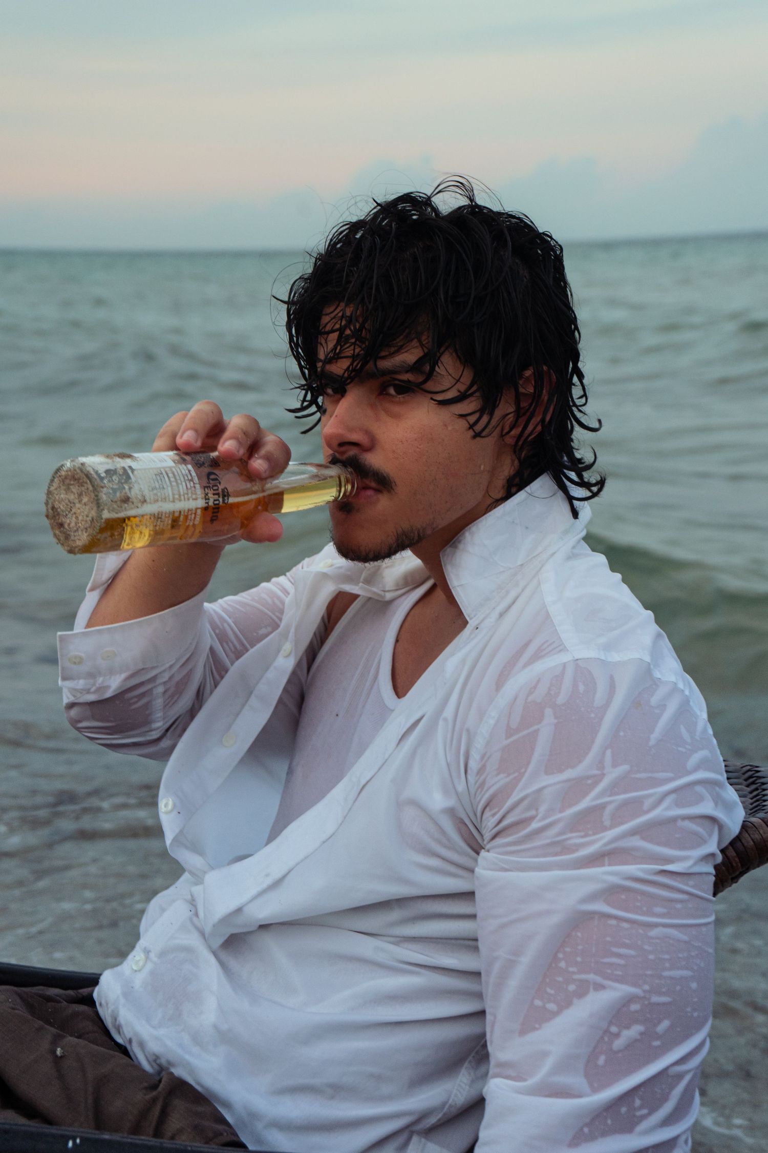 Young man drinking from a bottle on the beach wearing a wet white shirt at sunset.
