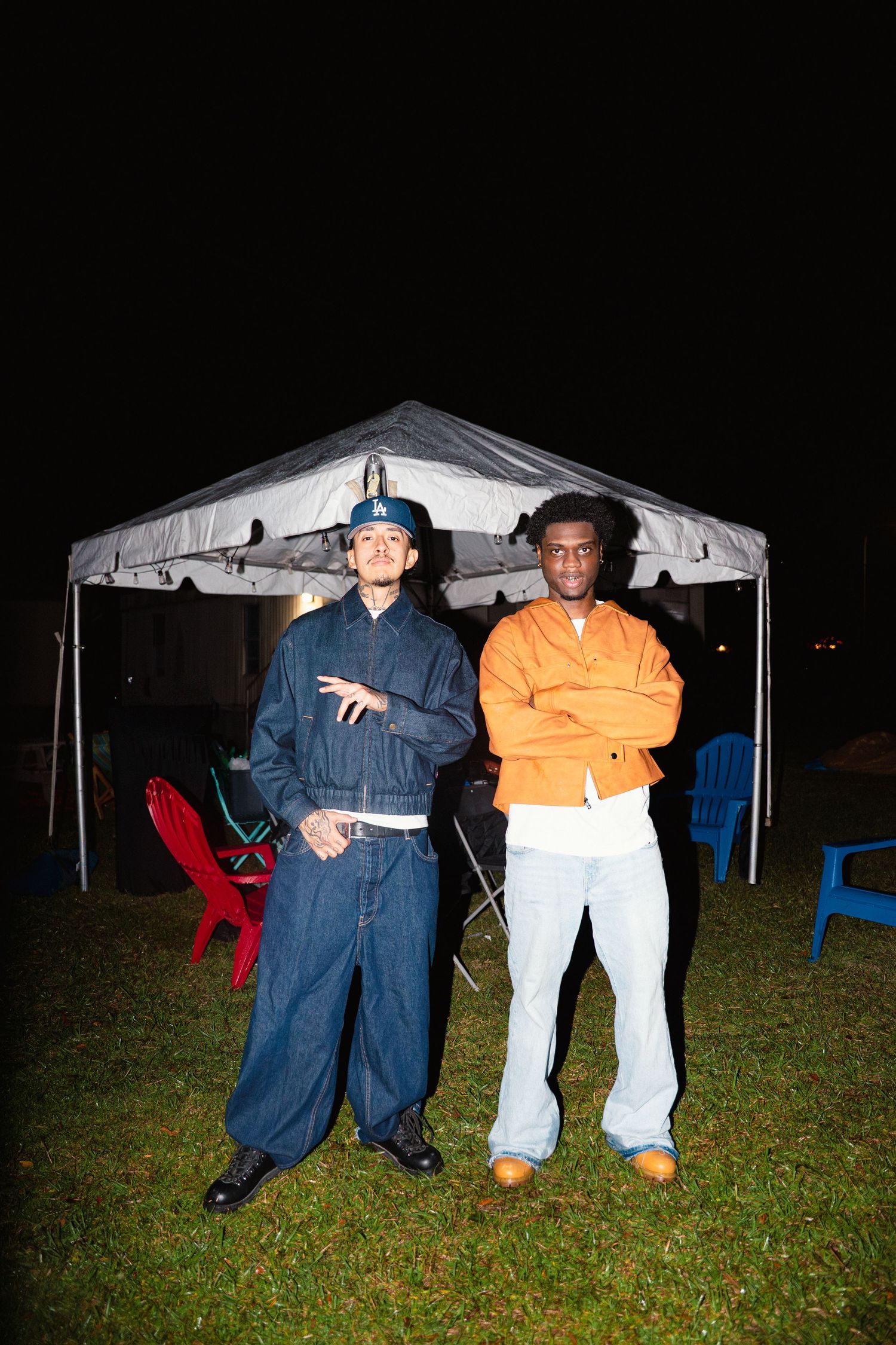 Two men posing outdoors at night near a tent, one in denim and one in an orange jacket.