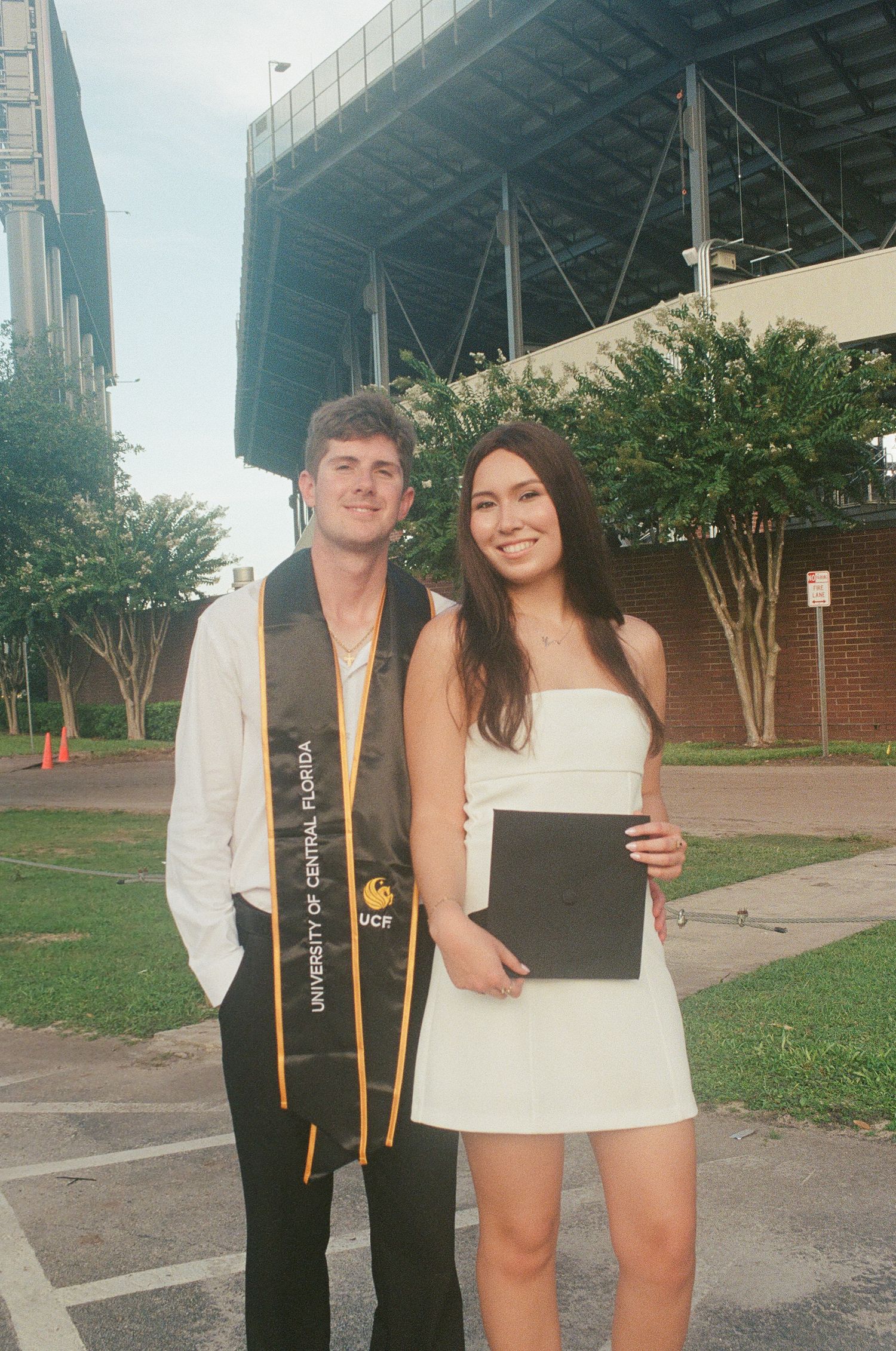 Two UCF graduates in stoles pose together outdoors holding diploma after ceremony.