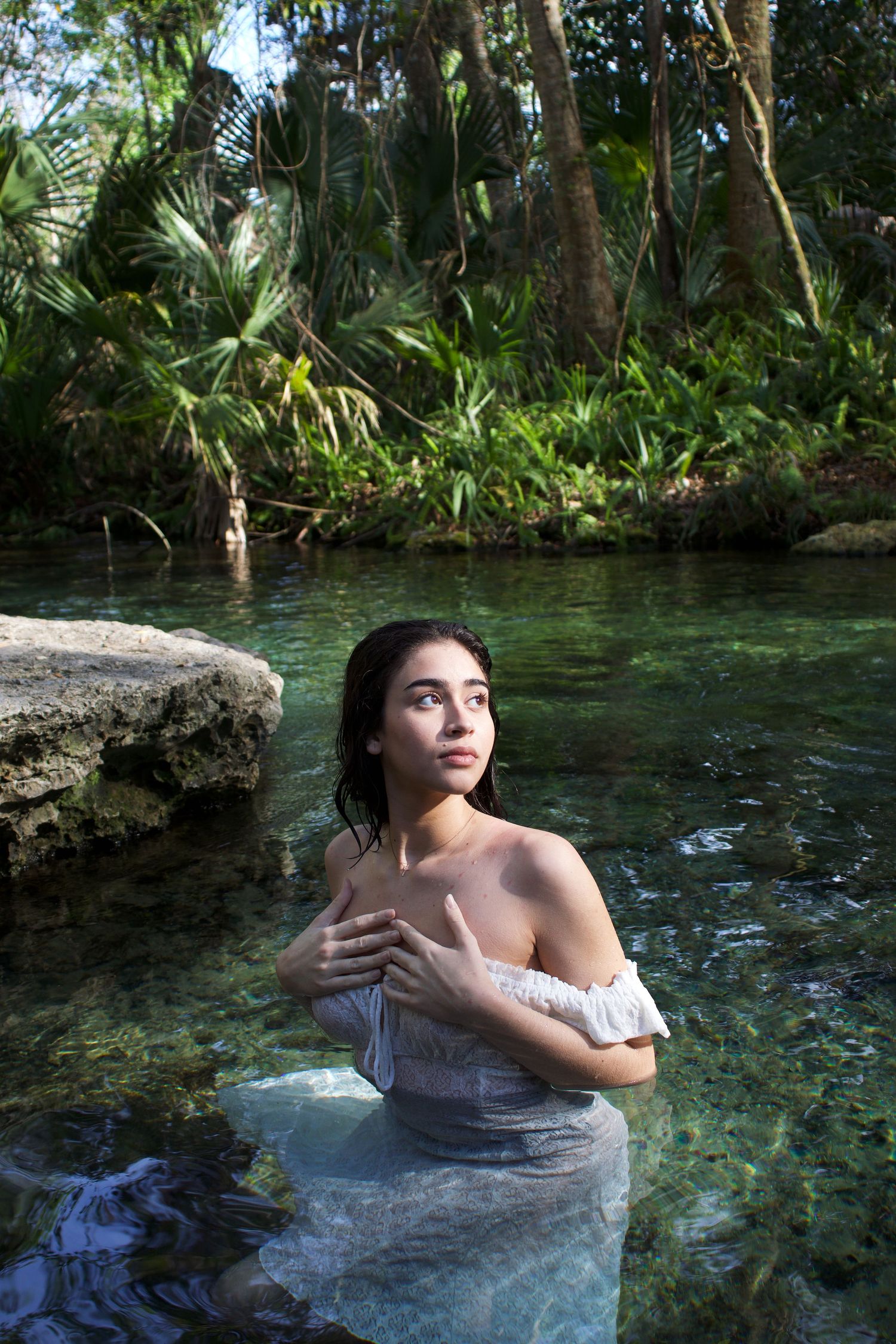 Young woman in white off-shoulder dress standing in clear natural spring water.
