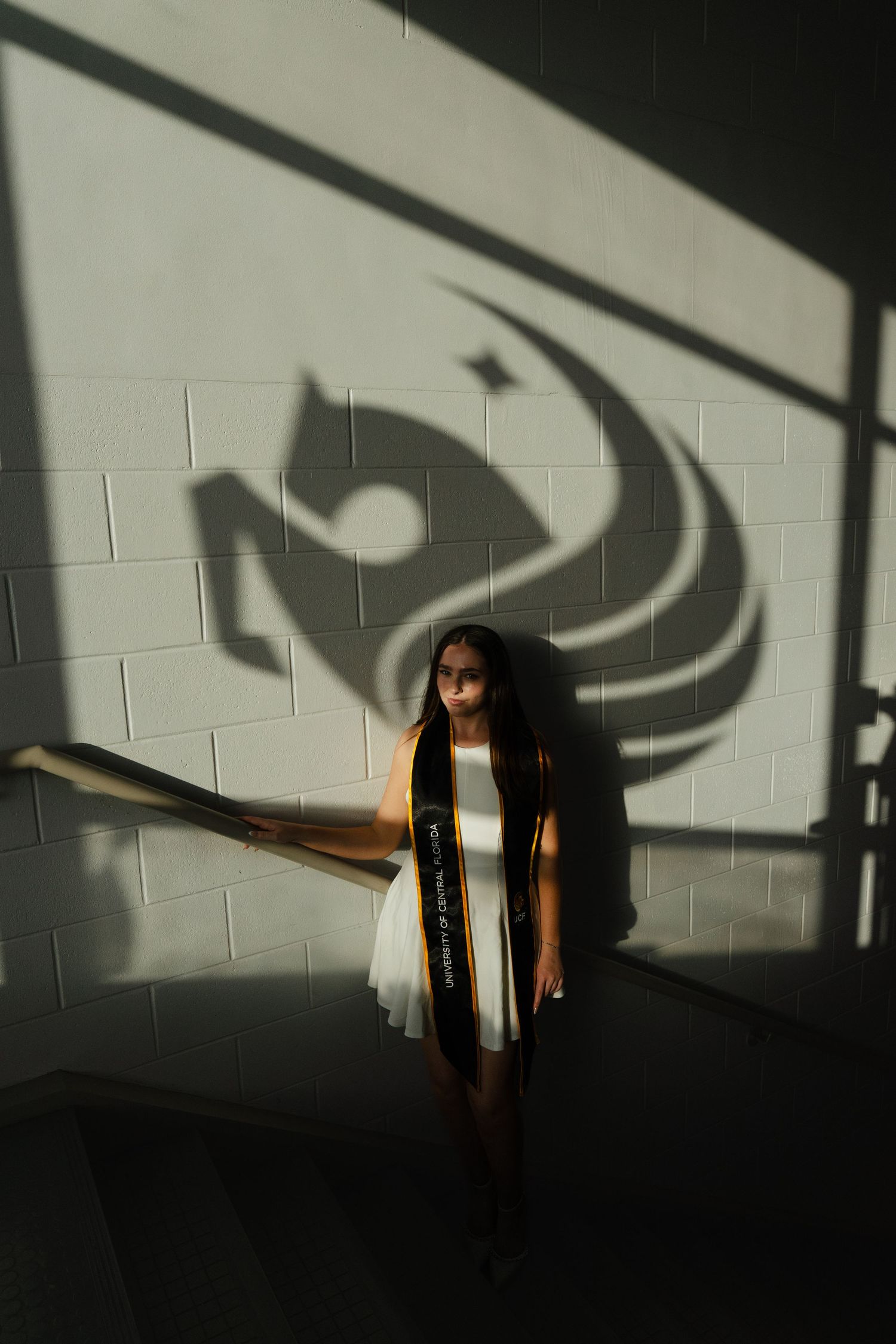 Graduate in white dress and black sash poses before dramatic shadow of school mascot on brick wall.