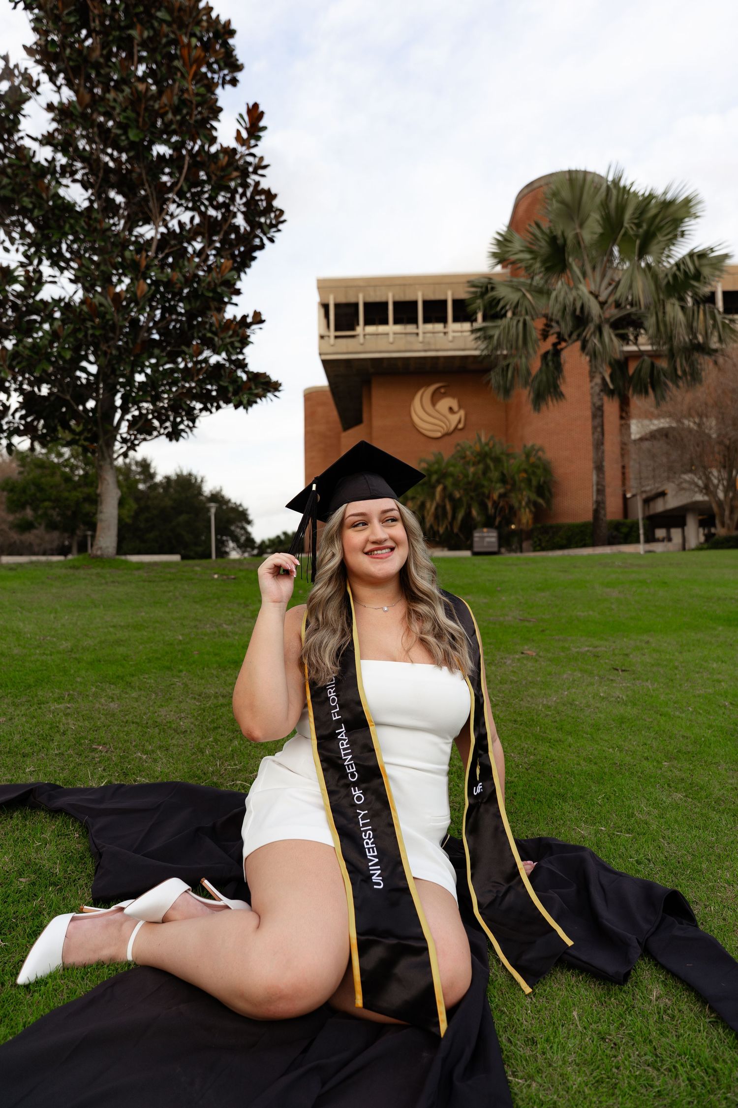 UCF graduate sits on grass in white dress with cap and University of Central Florida sash.