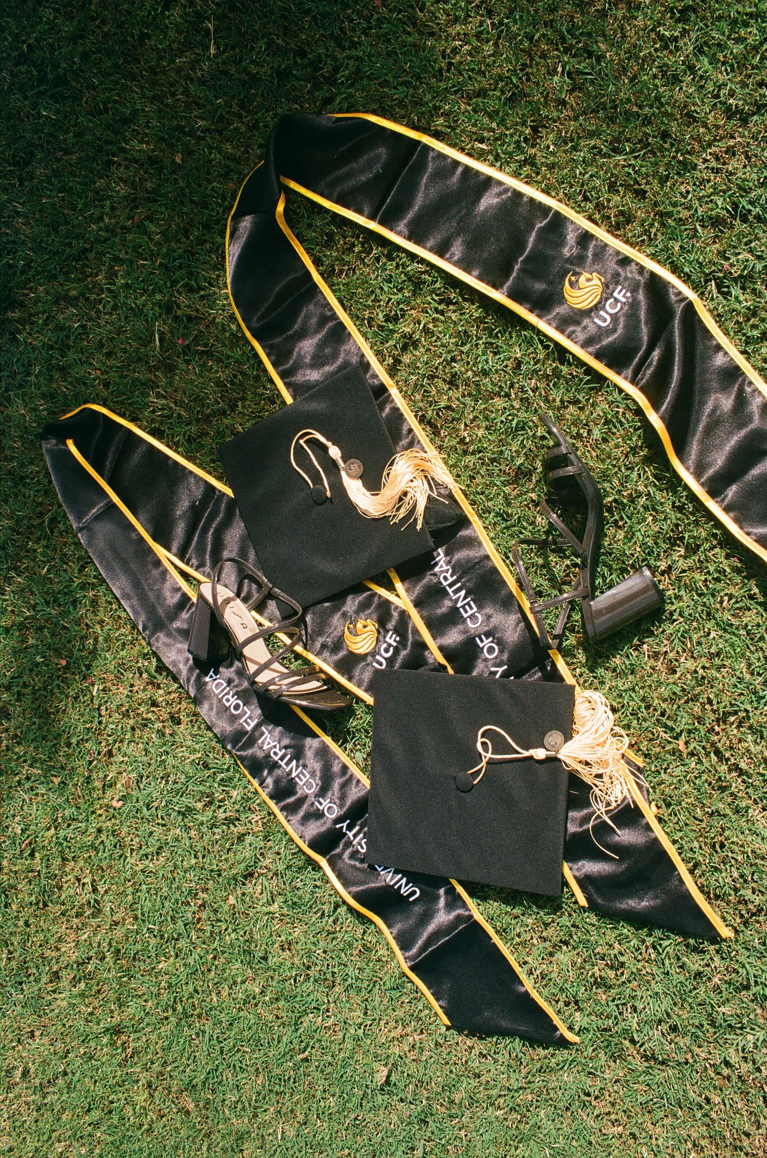 Black and gold graduation cap, tassel, and sash laid on green grass for a university commencement ceremony.